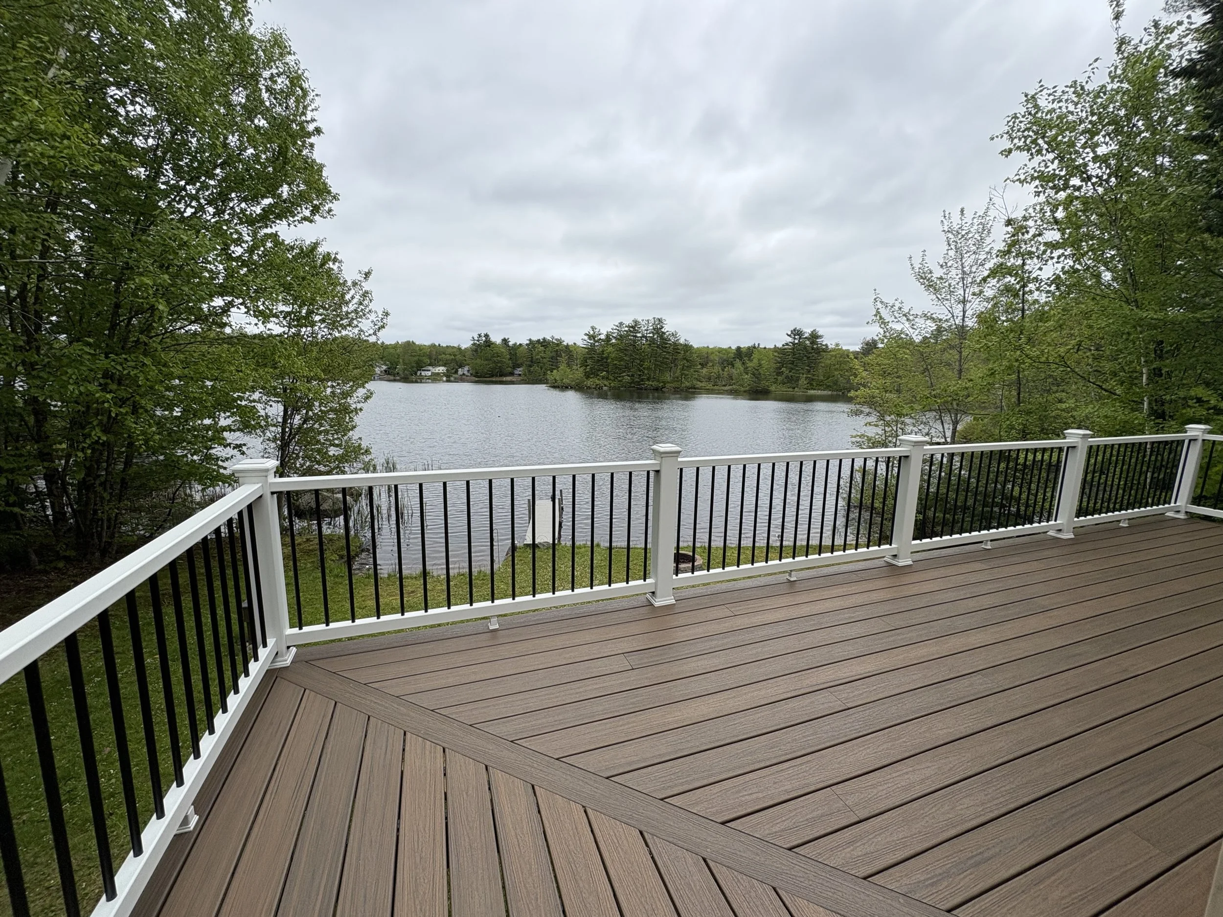 View of a lake from a wooden deck with a white and black railing, surrounded by trees and cloudy sky.