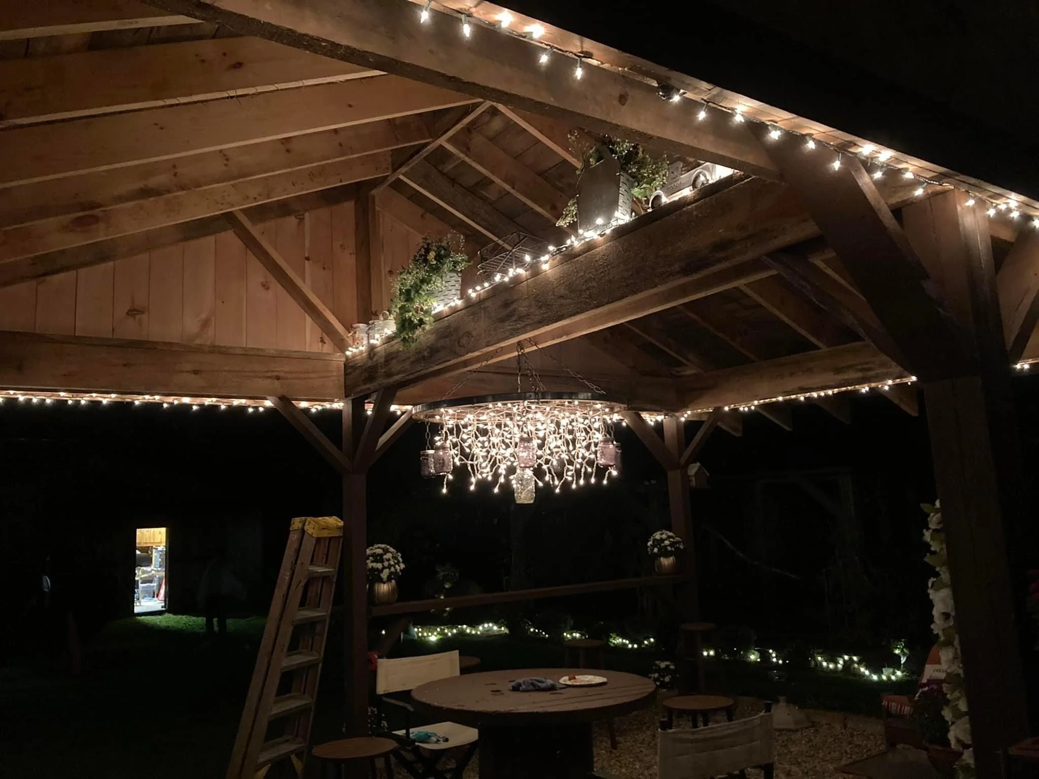 A wooden outdoor pavilion decorated with string lights, a chandelier, potted plants, and flowers, set up for an evening gathering.