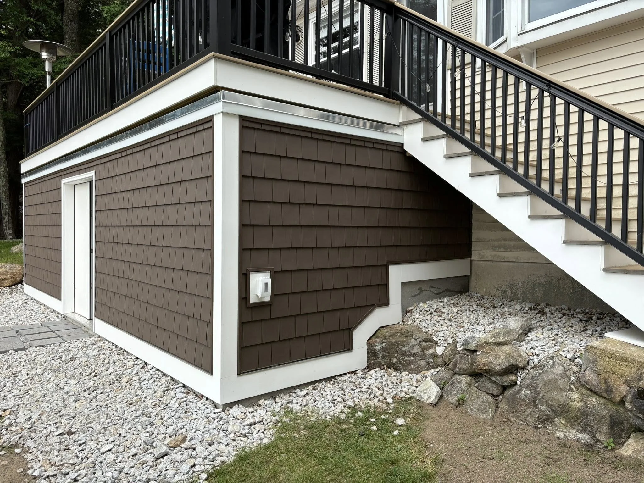 Exterior view of a house with brown siding, a staircase with black railings, and a small enclosed area at the bottom of the stairs, surrounded by rocks and gravel.