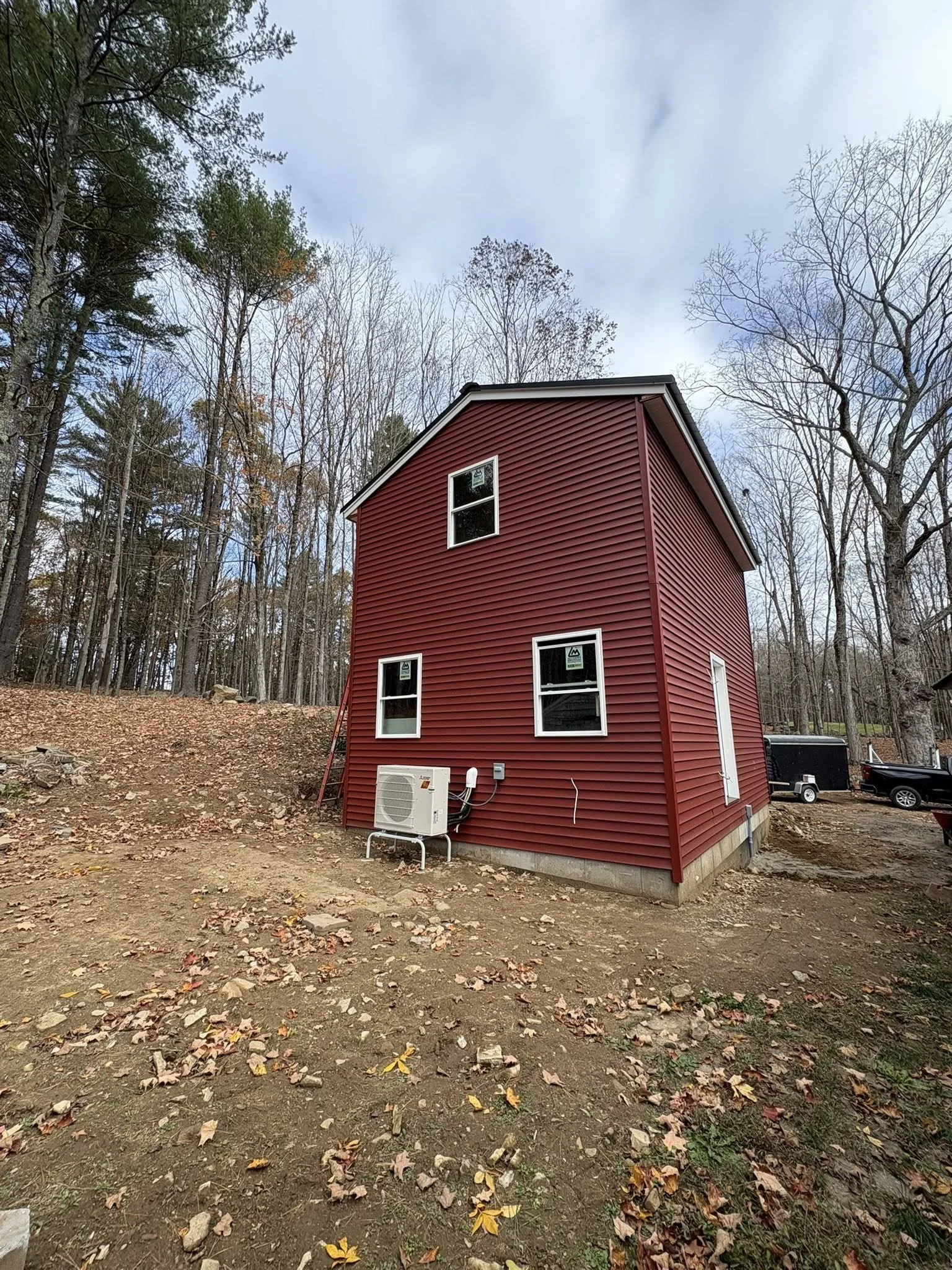 A new two-story house with red siding, three windows on the visible side, and an exterior air conditioning unit outside on a dirt yard surrounded by trees.