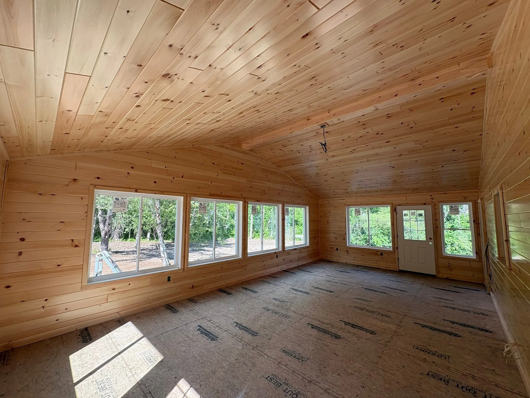 Interior view of a room under construction with wood-paneled walls and ceiling, windows overlooking a wooded area, and a floor covered with insulation material.