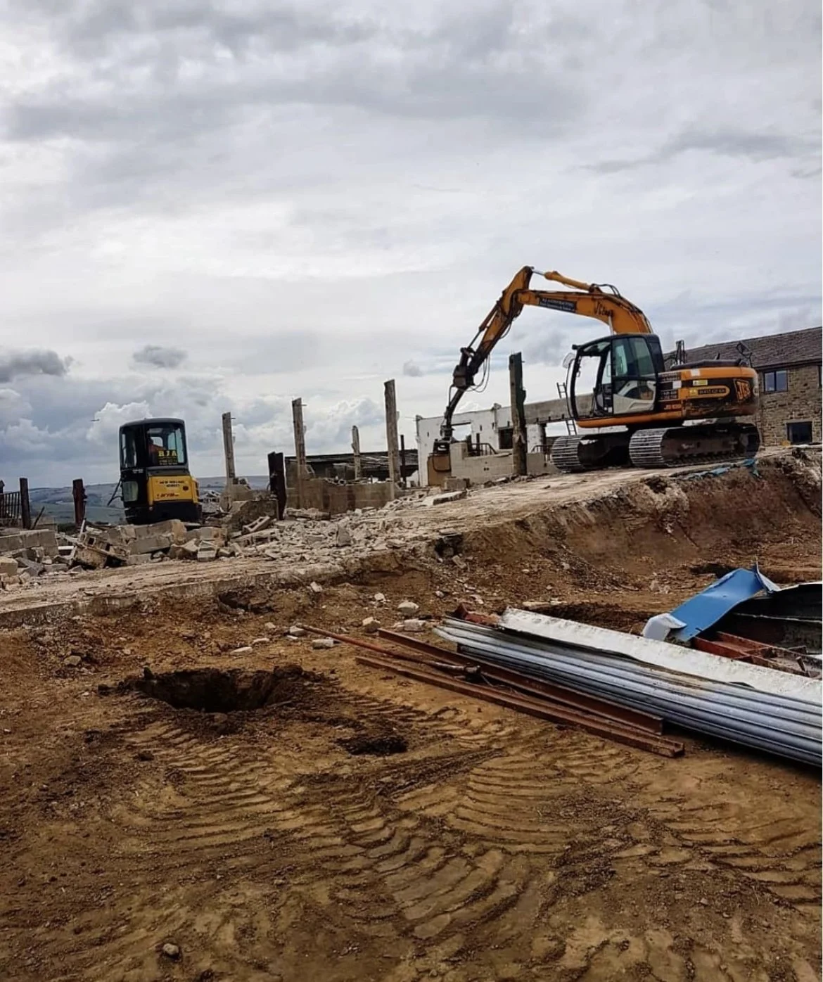 A construction site with an excavator and a small bulldozer working on a building foundation, with scattered construction materials and a cloudy sky in the background.