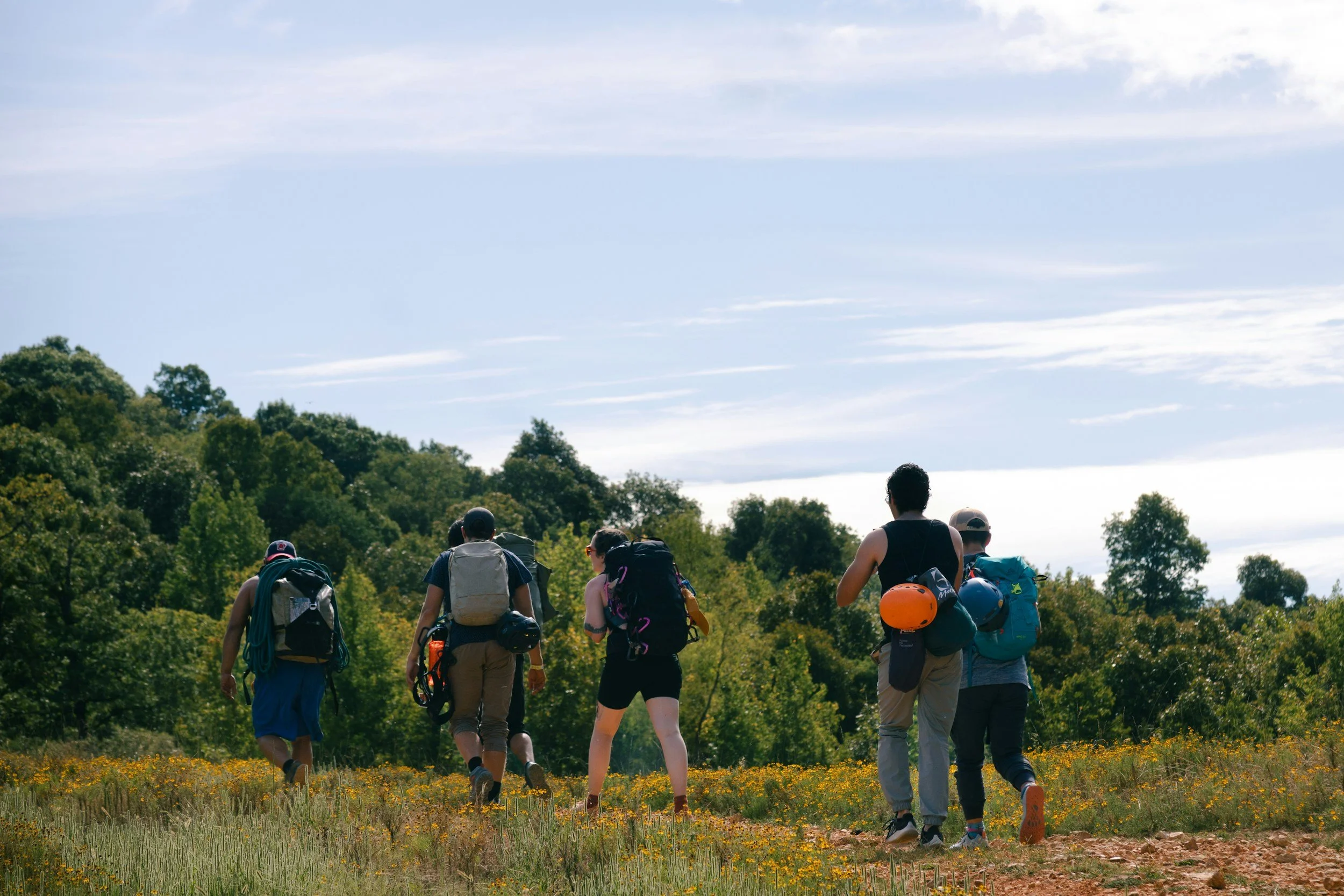 A group of hikers with backpacks walking through a grassy field with yellow flowers, surrounded by trees and under a partly cloudy sky.