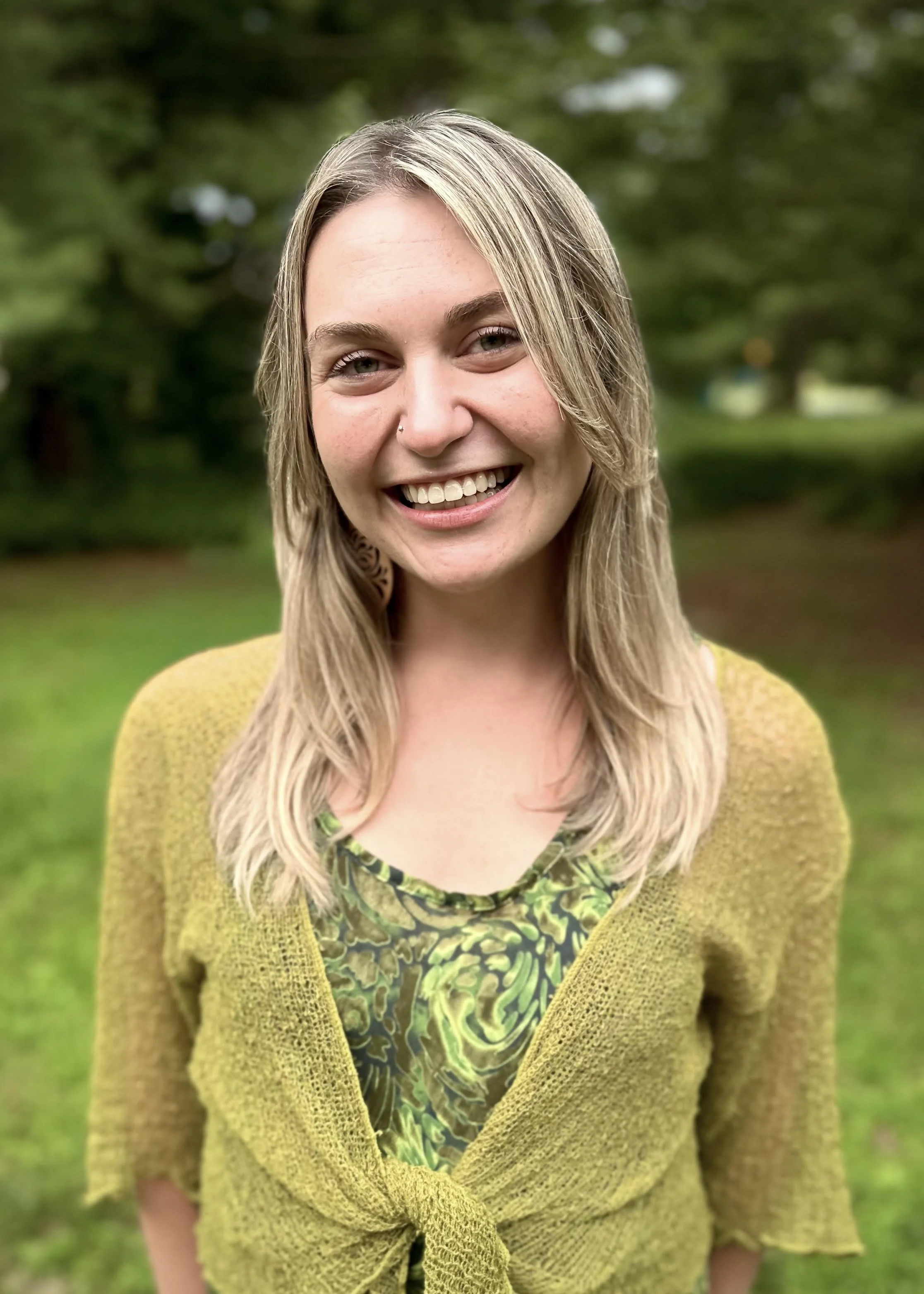 A smiling woman with long blonde hair, green eyes, and a nose piercing, wearing a yellow knit cardigan over a patterned green and black dress, standing outdoors with a blurred green background.