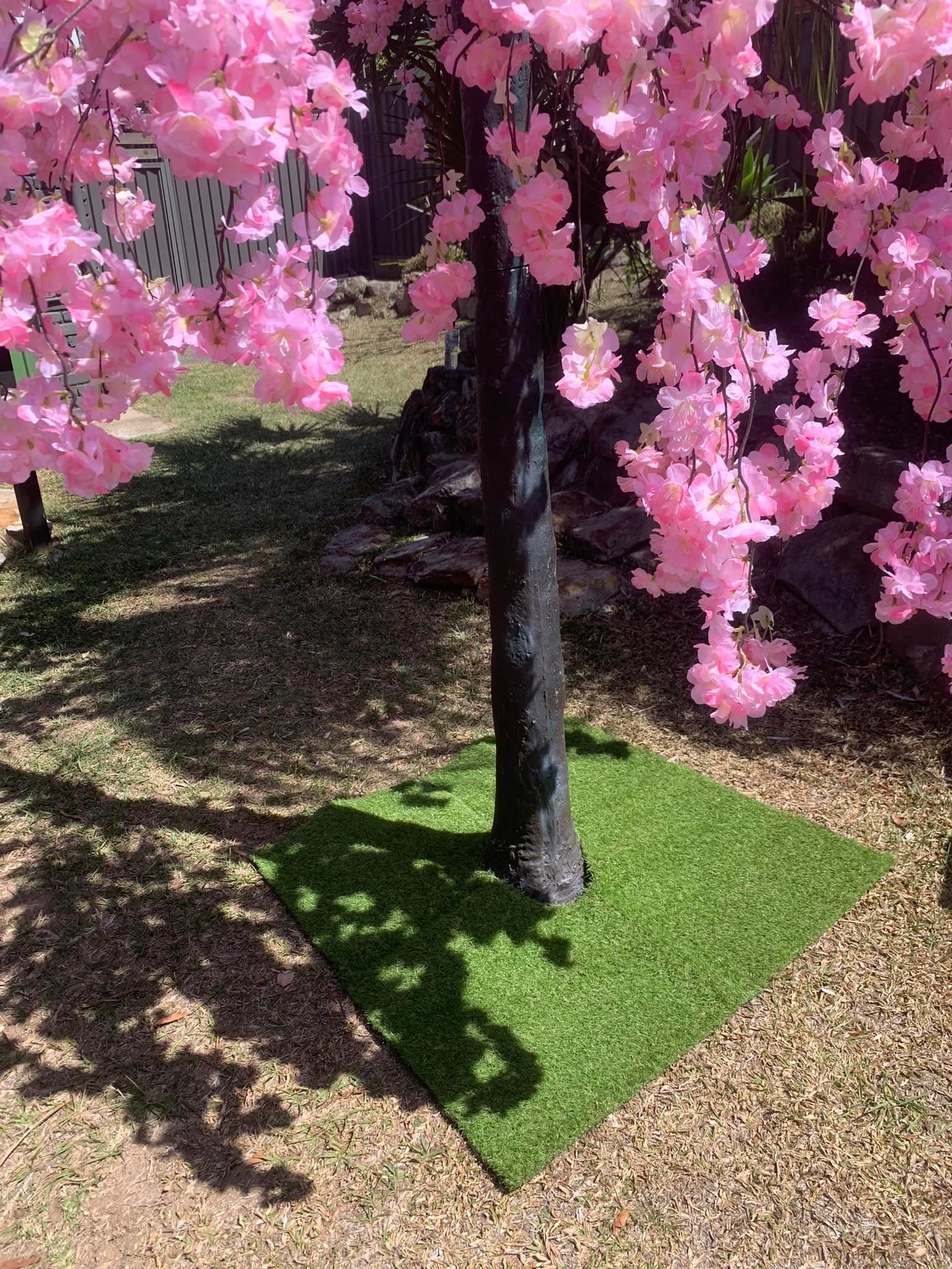 Artificial pink cherry blossom tree with a black trunk on a square of green artificial grass in a garden setting.