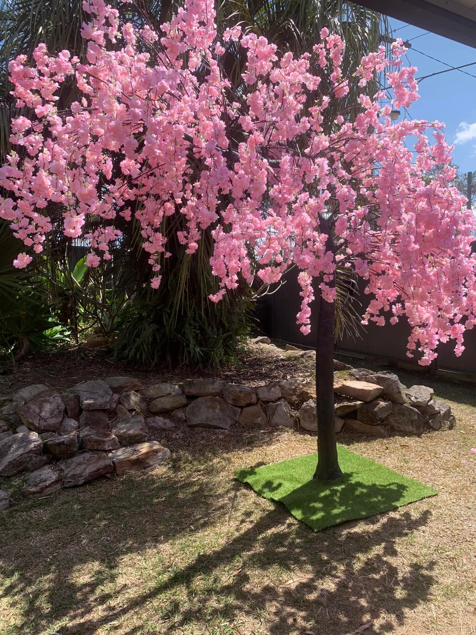 Artificial cherry blossom tree with pink flowers in a garden setting, surrounded by rocks and greenery.