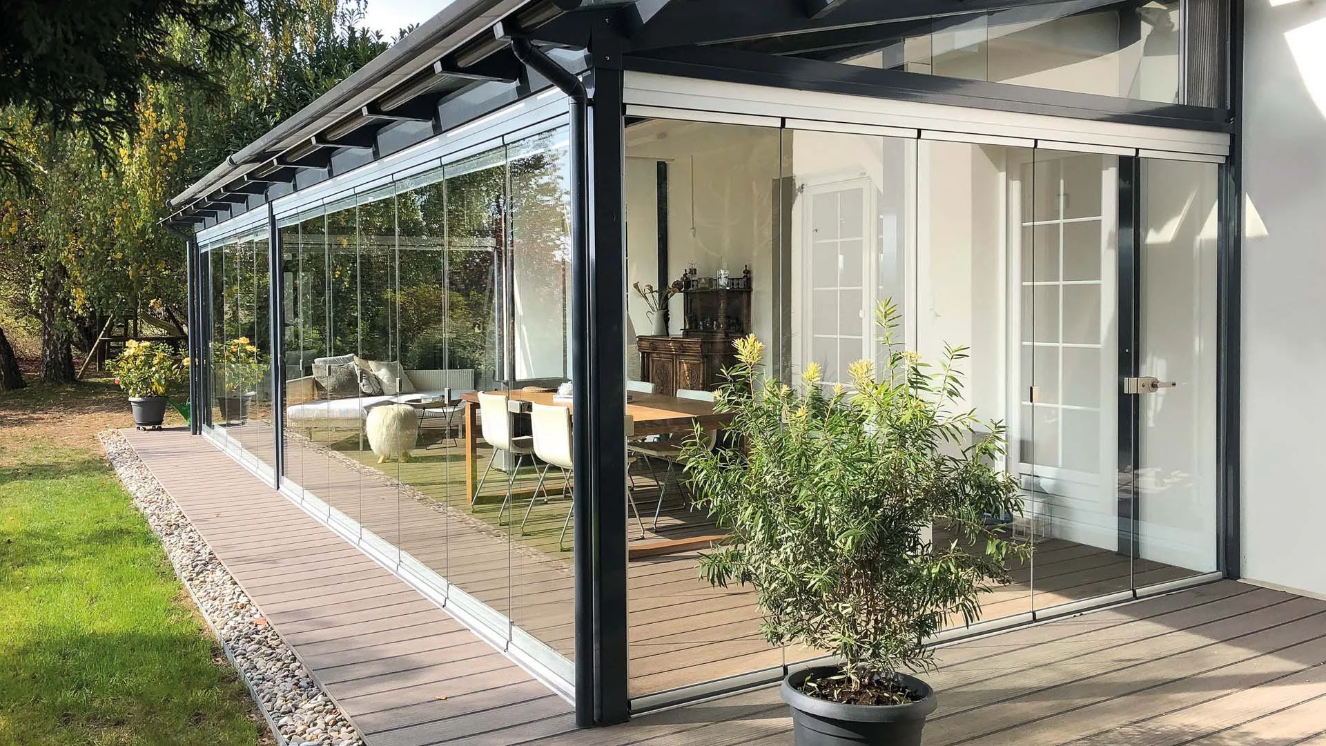 A modern glass-enclosed patio attached to a house with a wooden deck, potted plants, and a view of the interior dining and living area.