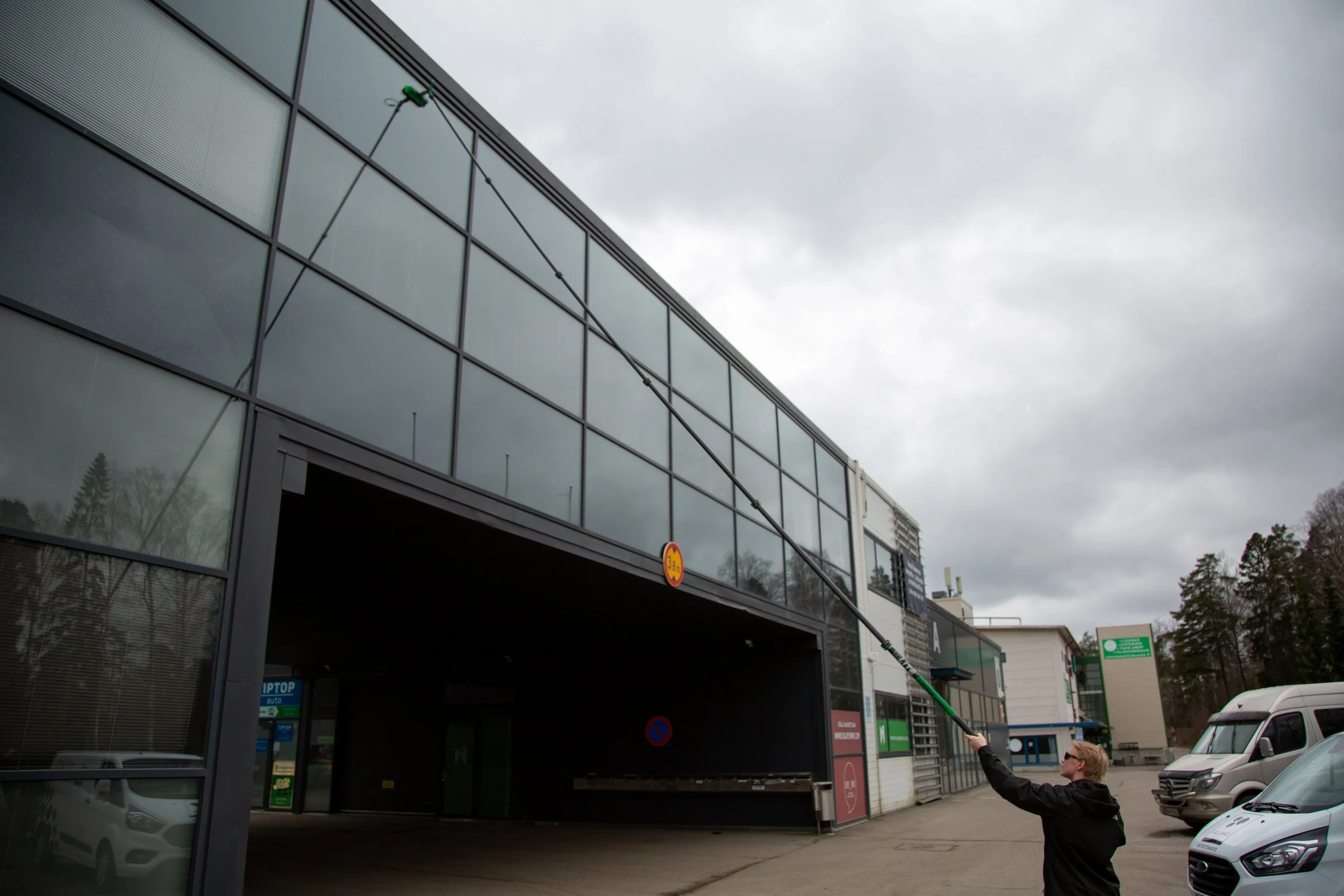 Person cleaning the exterior glass of a modern commercial building with a long-handled tool, parked cars visible on a cloudy day.