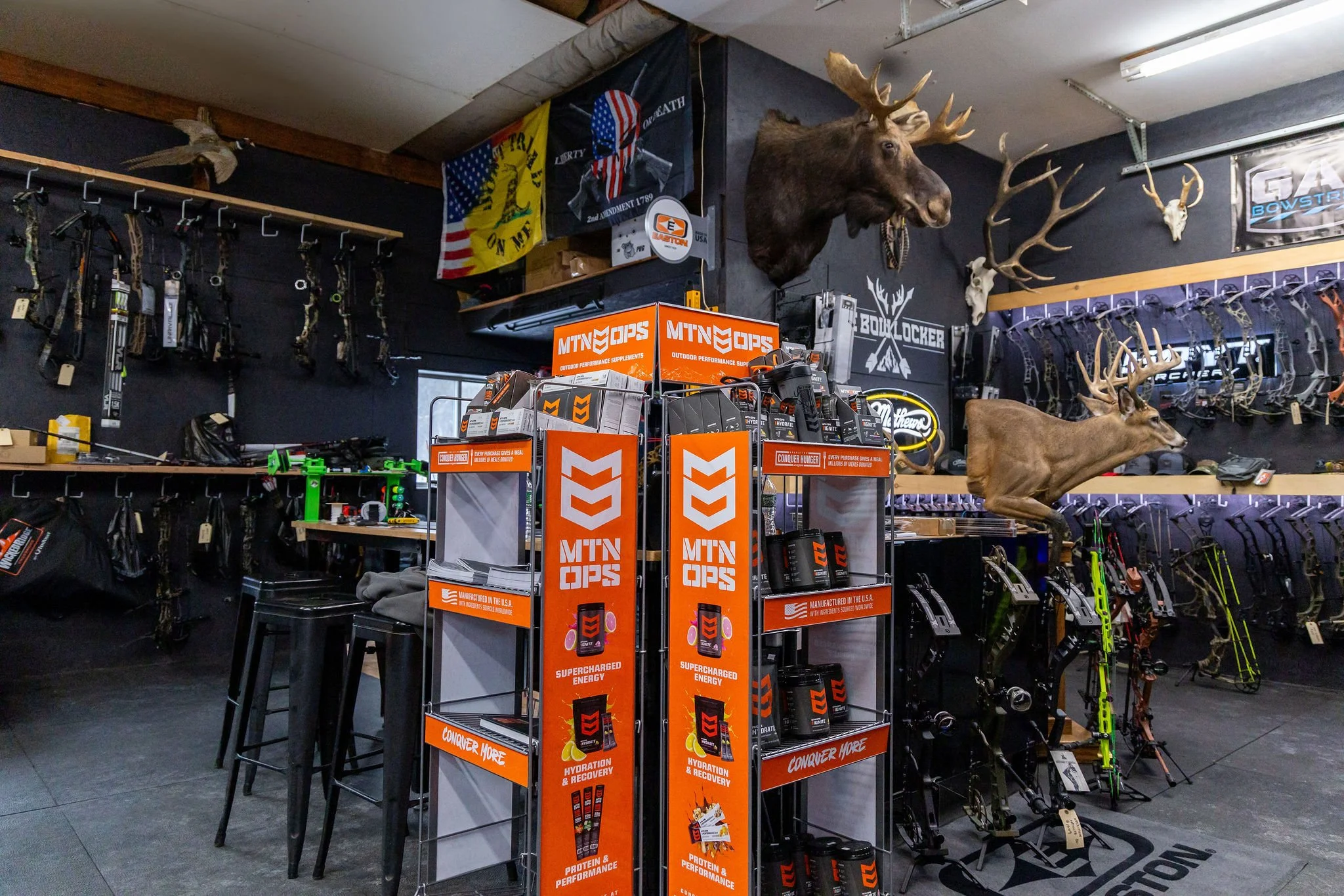 Inside a hunting or outdoor sporting store, with deer heads mounted on the wall, hunting bows displayed on the wall, and a display stand for mountain ops energy supplements in the center.