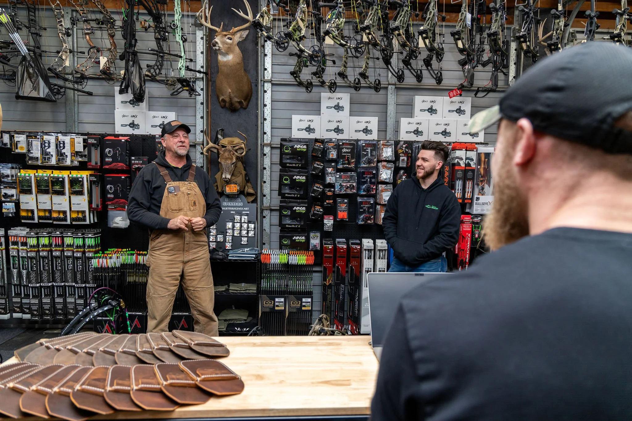 A man in brown overalls and a black cap is speaking to two men in a hunting or sporting goods store, with hunting gear like bows, arrows, and mounted animal heads on the wall behind them.