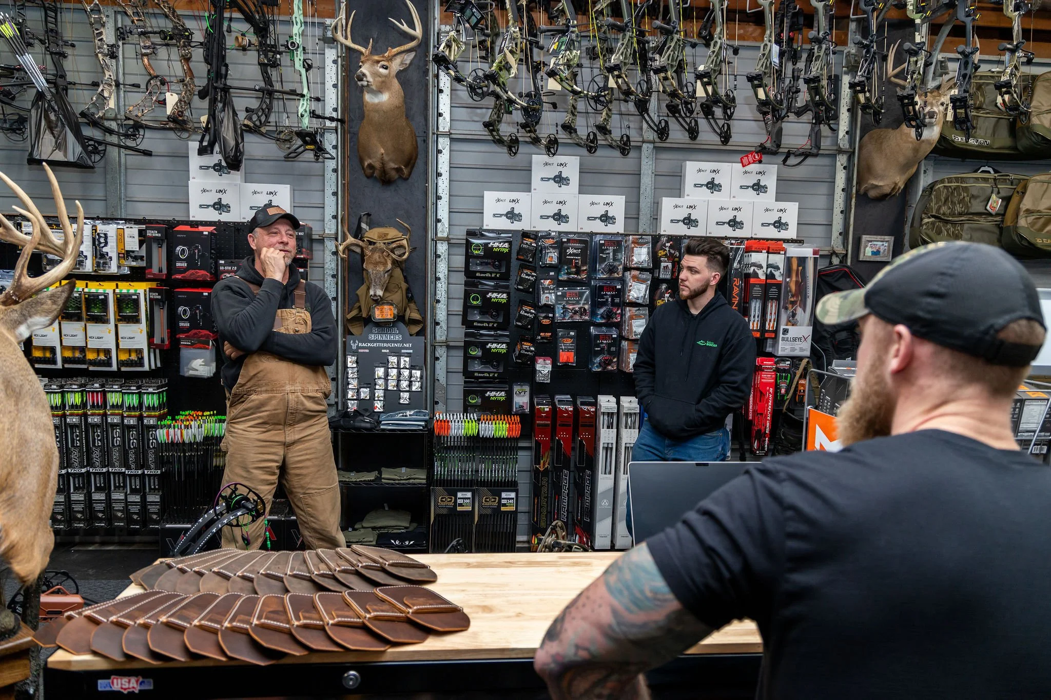 Inside a hunting store with mounted deer heads and gear displayed, three men are having a conversation. One man is wearing overalls, another in a black hoodie with hands in pockets, and the third, with tattoos and a black cap, is sitting at a table.