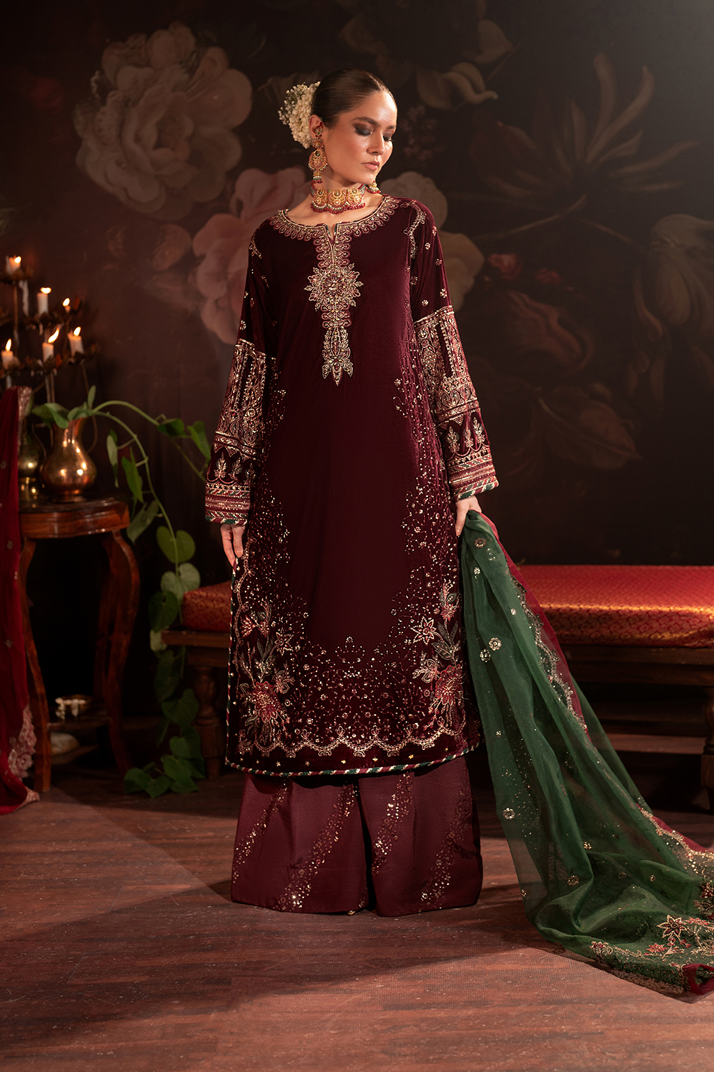 Woman wearing an ornate maroon traditional dress with gold embroidery, paired with large gold jewelry, standing indoors with dark floral wallpaper background and lit candles.