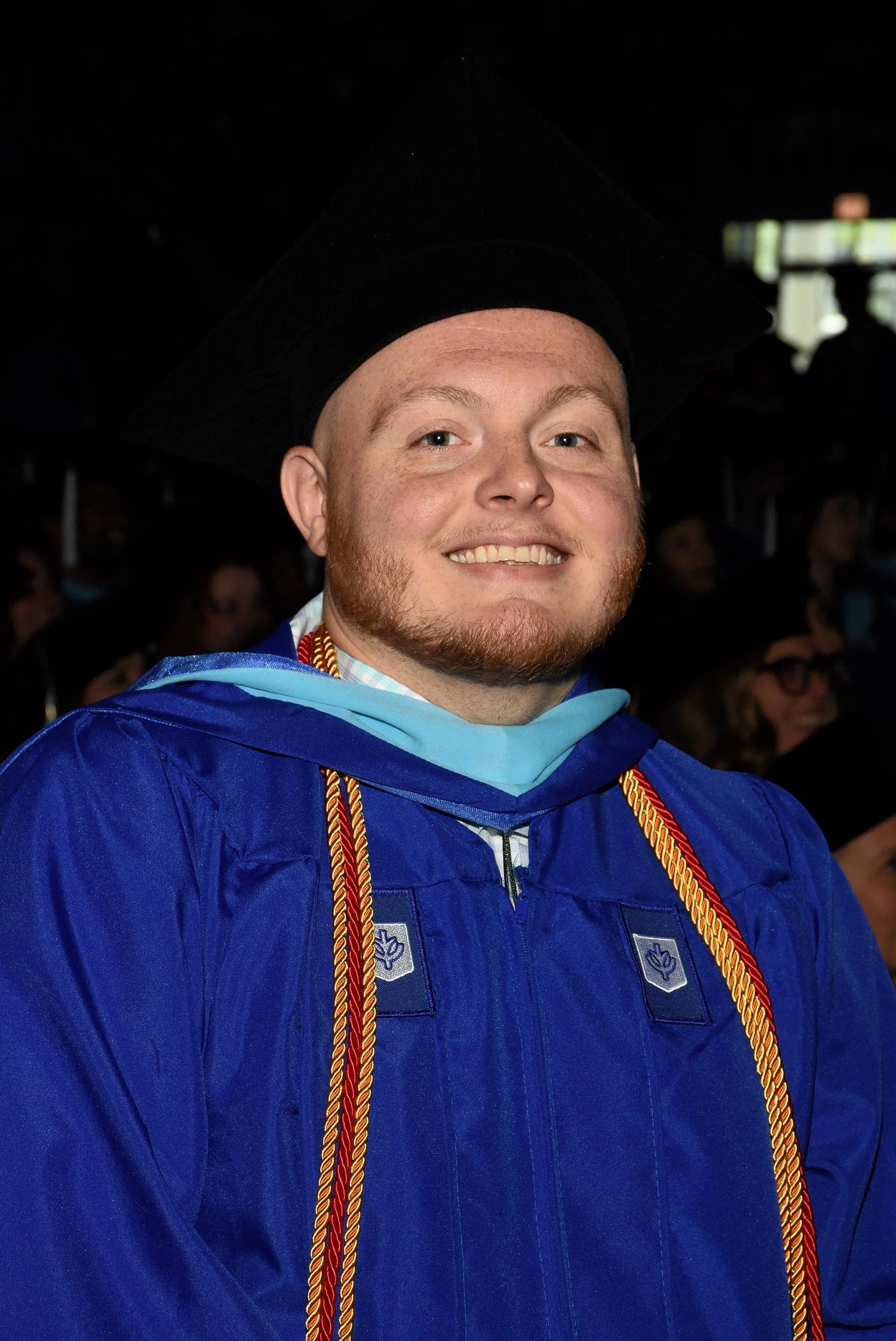 A young man with light skin, short red hair and beard, smiling and wearing a blue graduation gown and cap with red and gold cords around his neck during a graduation ceremony.