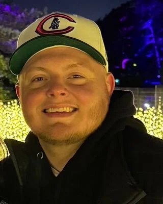 A smiling man wearing a baseball cap with the logo of the Cincinnati Reds, standing outdoors at night near a display of yellow lights.