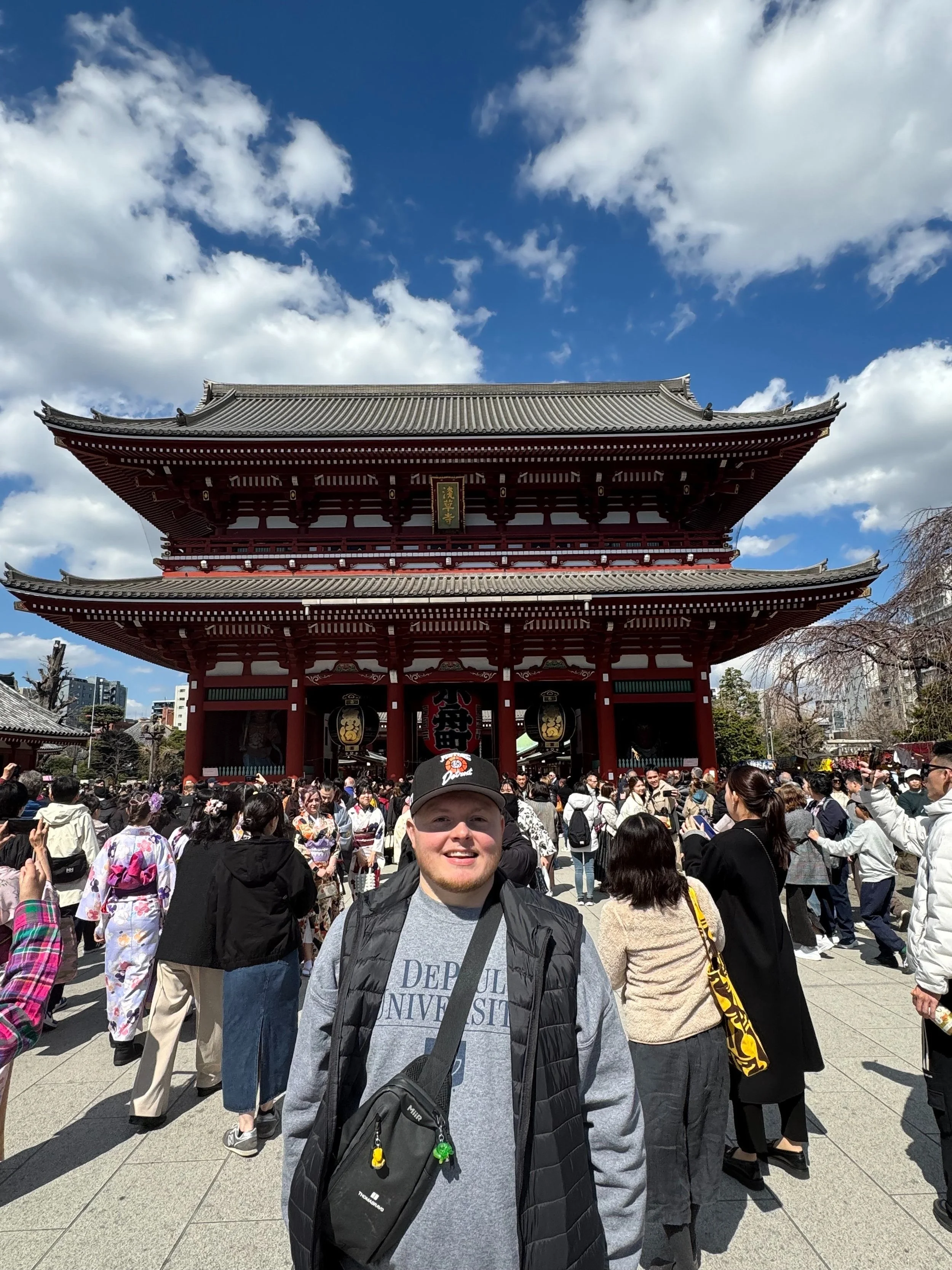 A smiling man with a gray sweatshirt, black jacket, and backpack standing in front of a large crowd at a traditional Japanese temple with a multi-tiered roof and a sign with Japanese characters, under a partly cloudy sky.