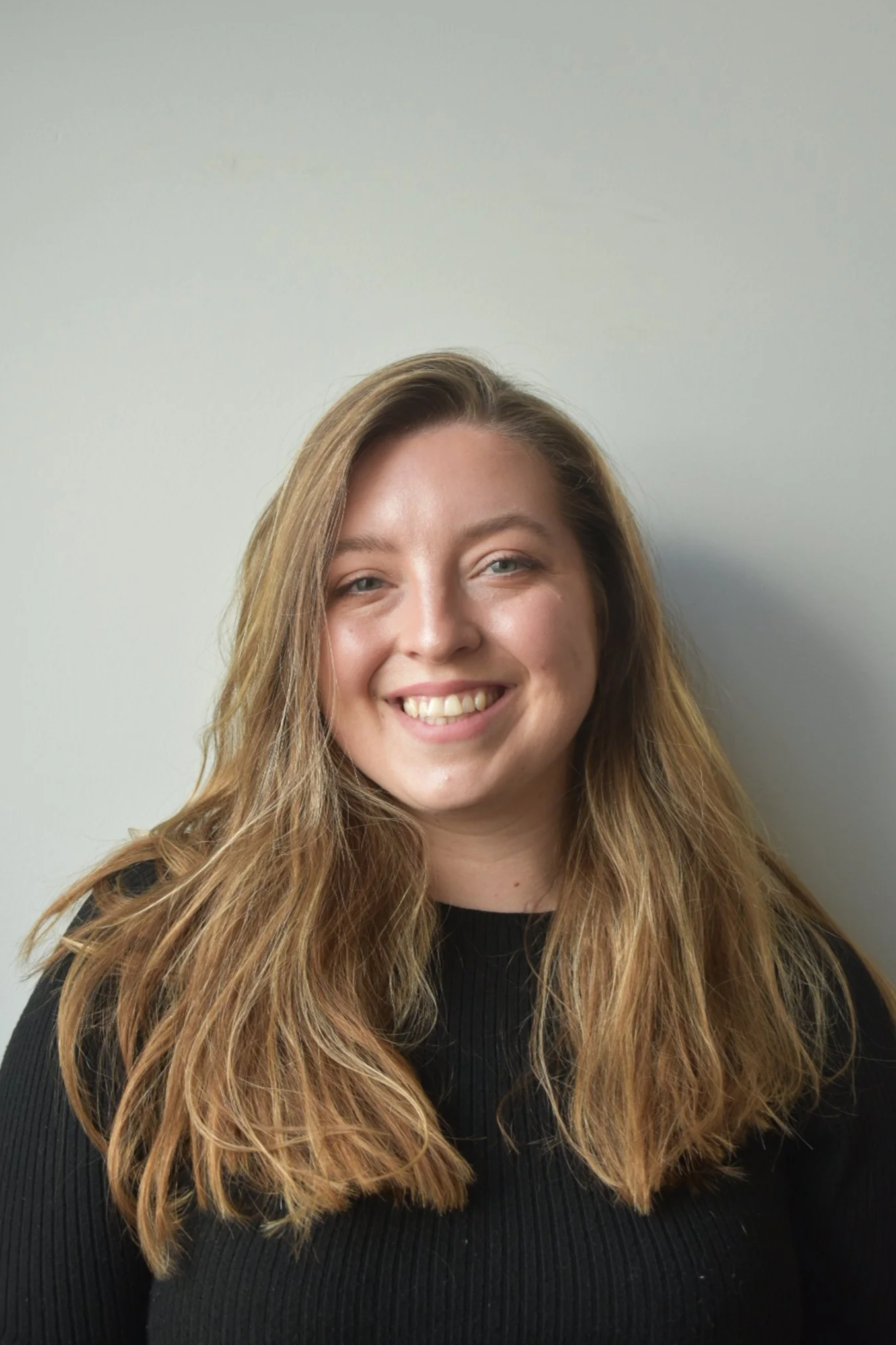 Portrait of a young woman with long, wavy brown hair, smiling, wearing a black top, against a plain white wall.