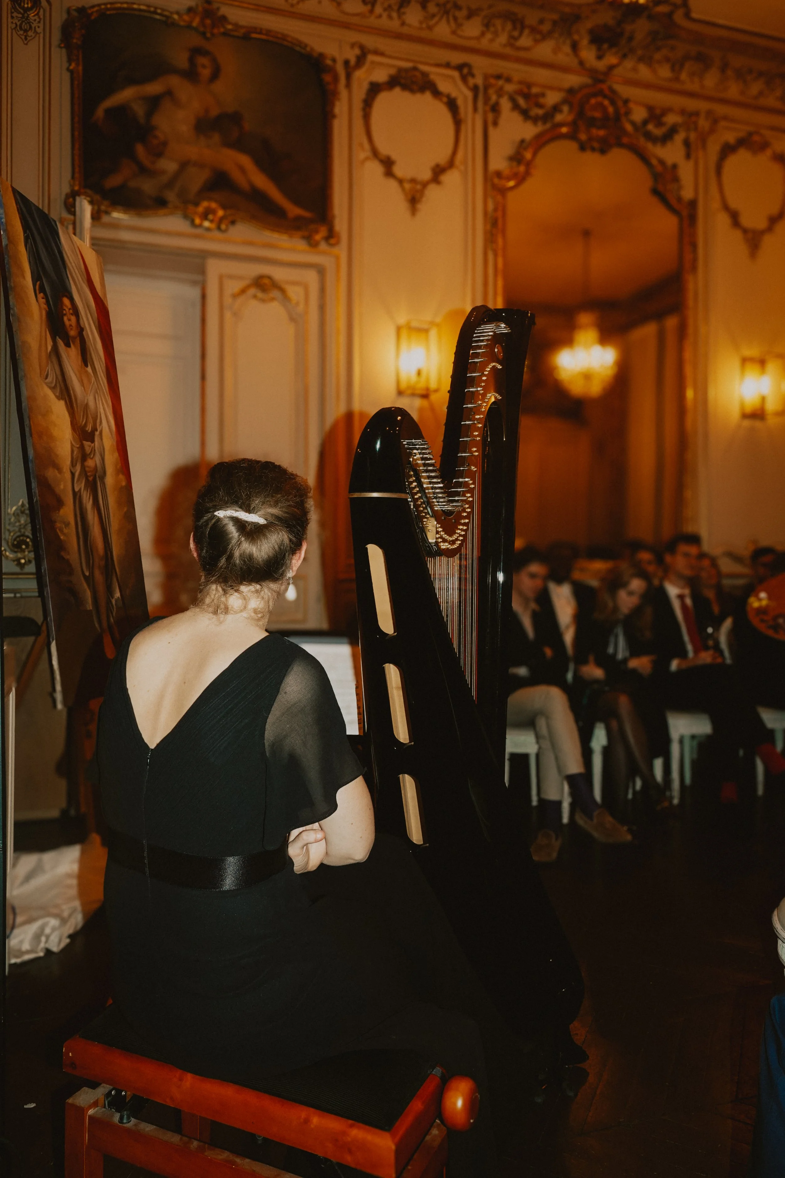 Une femme jouant du piano à queue devant un auditoire dans une salle ornée de dorures et d'œuvres d'art classiques.