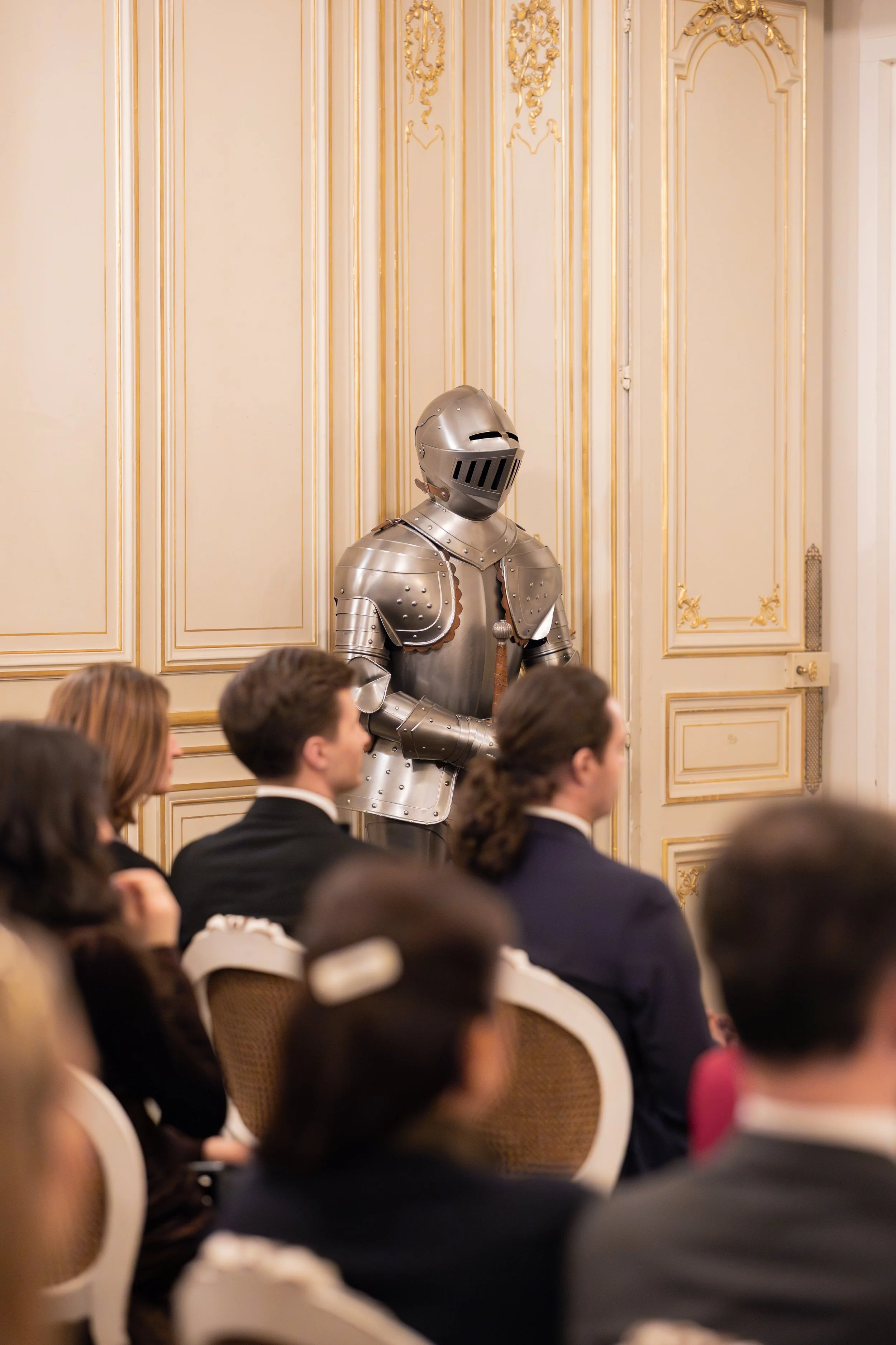 une personne en armure de chevalier en métal, assise dans une salle ornée de décorations en or, entourée de plusieurs personnes assises, lors d'un événement formel.