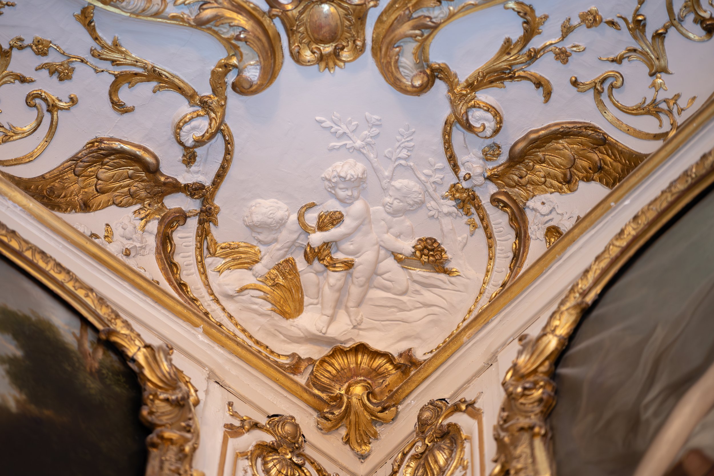 Détail d'une fresque ou sculpture ornementale au plafond illustrant des enfants, avec des éléments décoratifs dorés et blancs dans un style rococo.