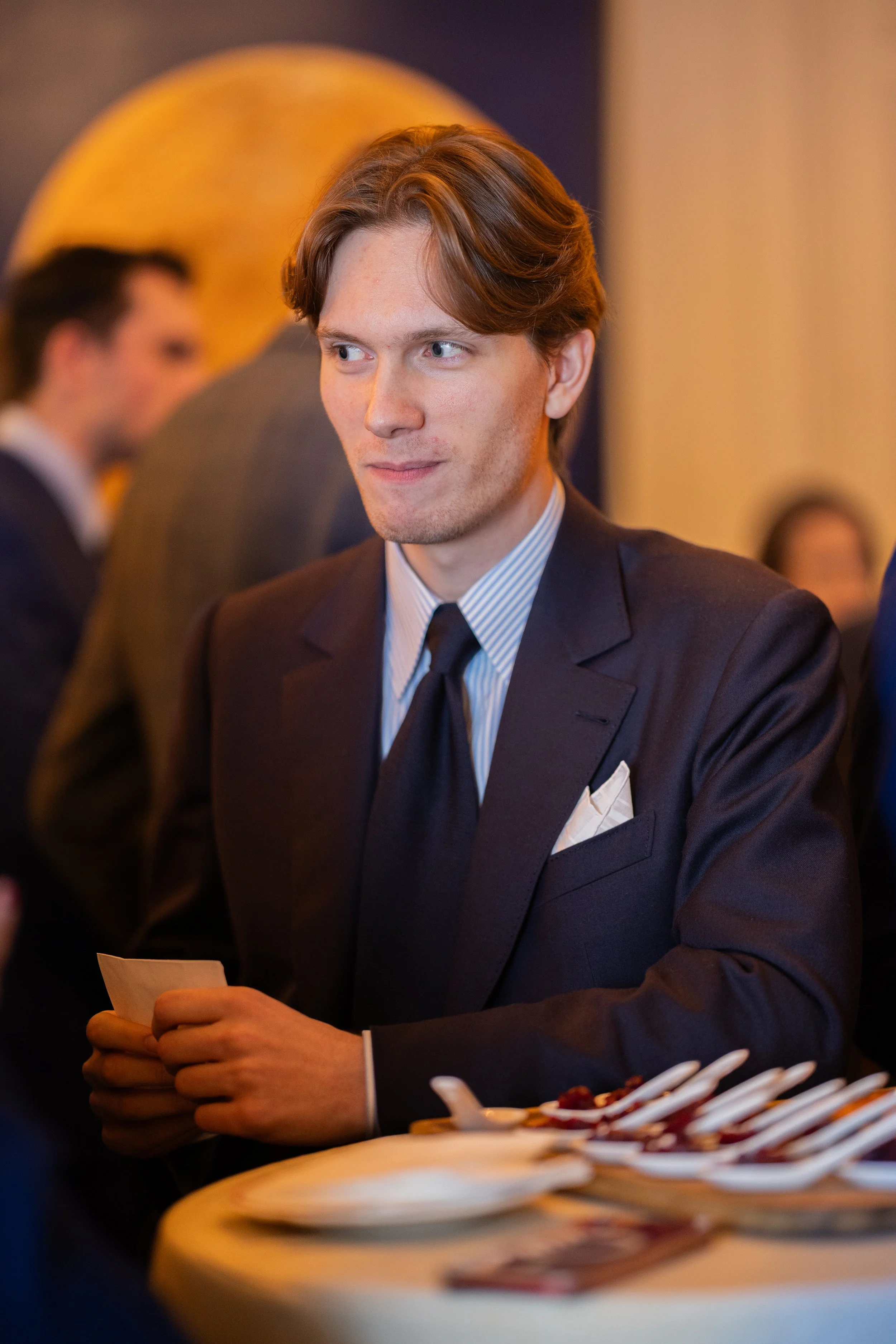 Jeune homme en costume à une réception, tenant un morceau de papier, avec une table de desserts en premier plan.
