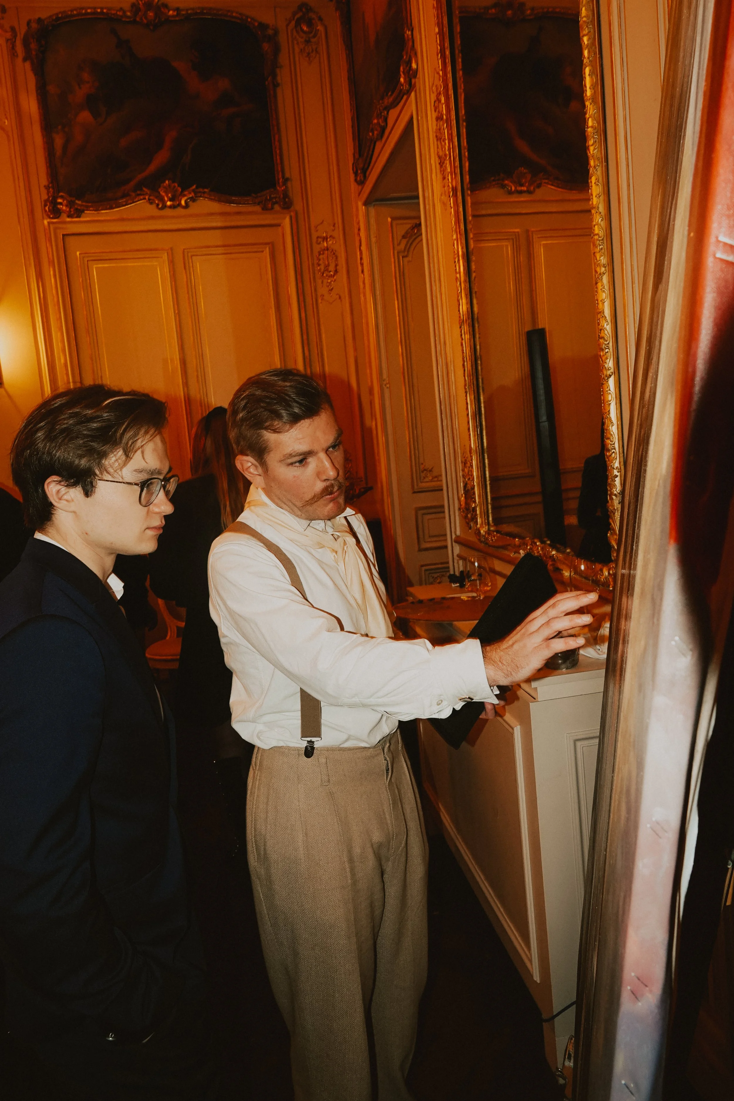 Deux hommes discutant devant un tableau dans une pièce élégante ornée de moulures dorées et de grands miroirs.