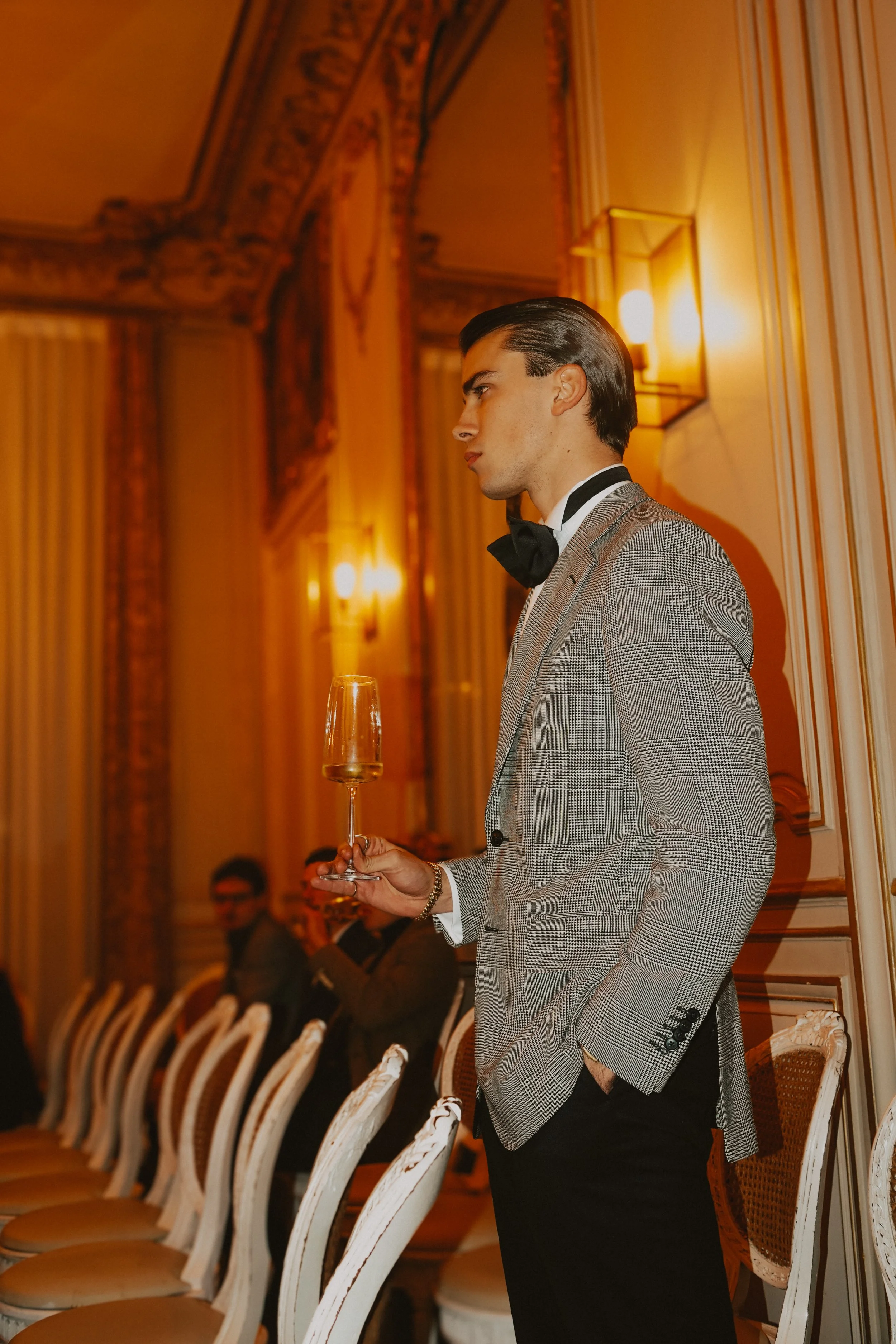 Jeune homme chic en costume à un événement élégant, tenant une flûte de champagne, dans une salle décorée de manière sophistiquée.
