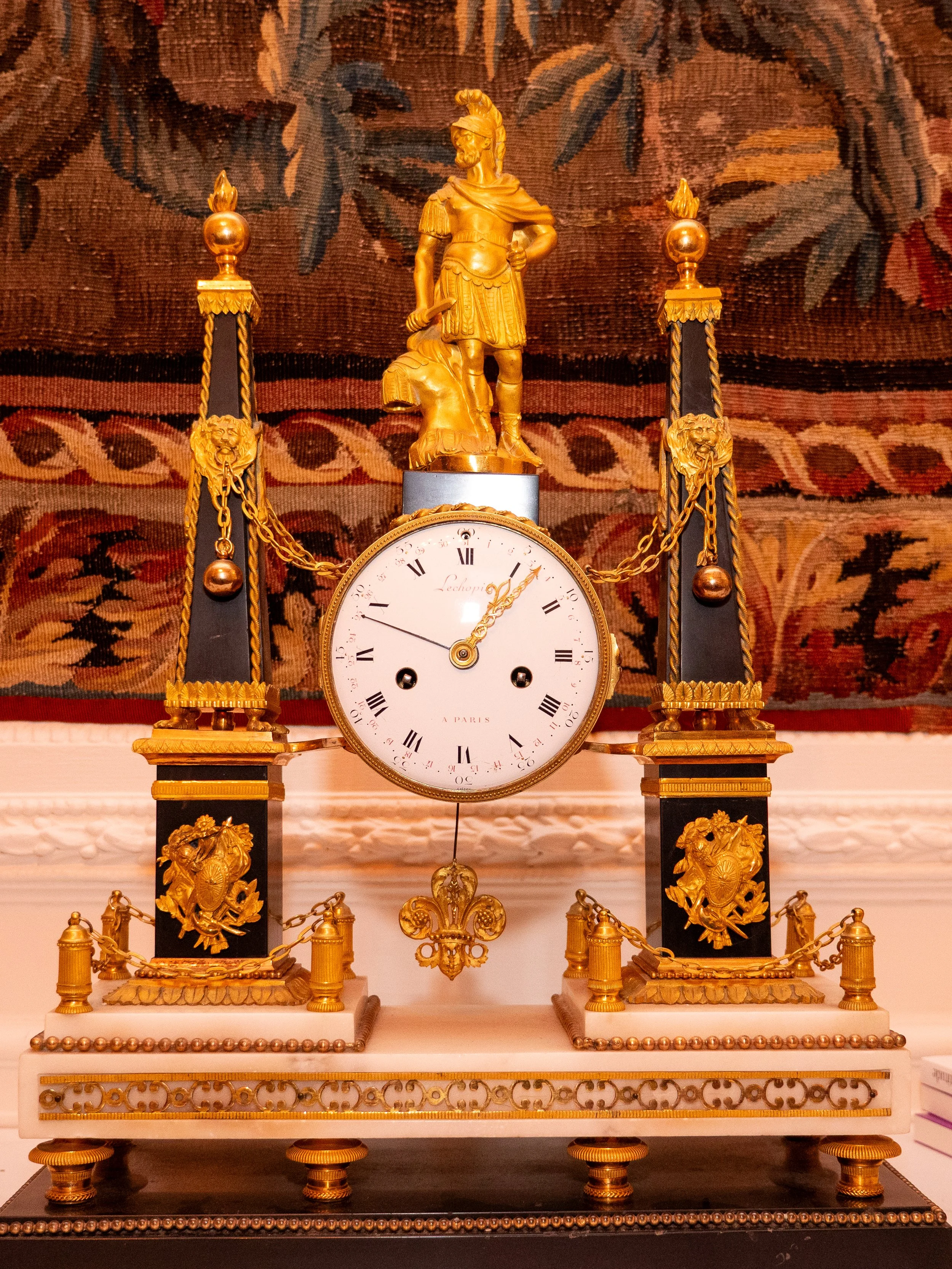 Une horloge ancienne avec des ornements dorés, une figure en haut et des décorations sur les côtés, posée sur une base en marbre blanc.