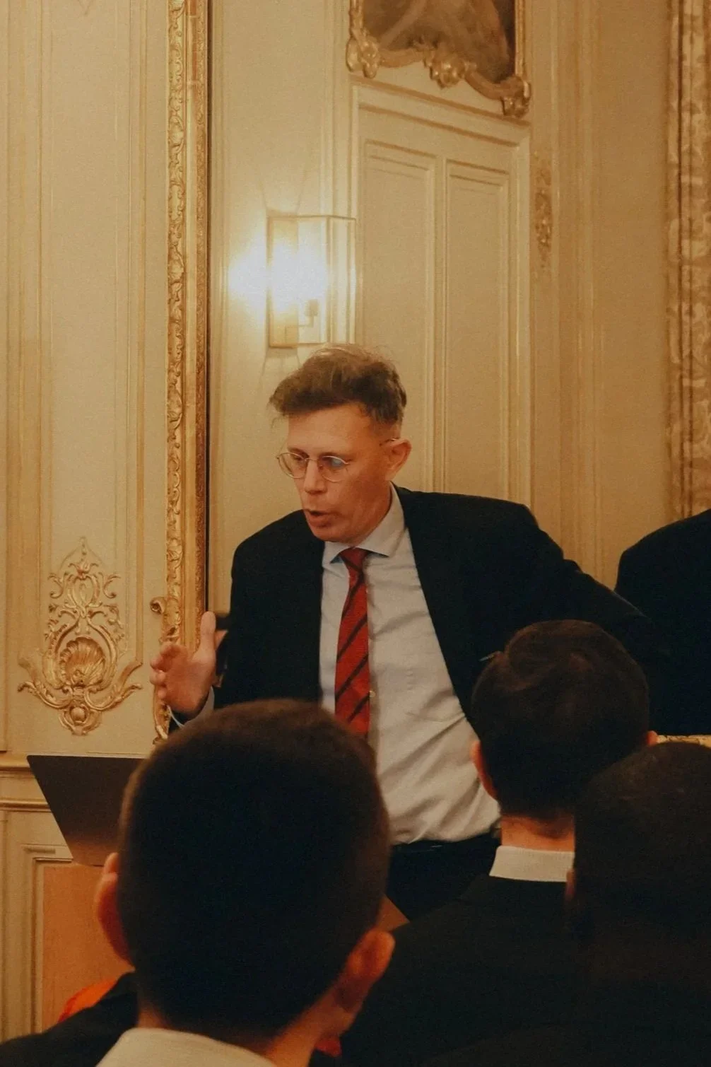 Un homme avec des lunettes et une cravate rouge parle lors d'une réunion ou d'une conférence dans une salle ornée de décorations classiques.