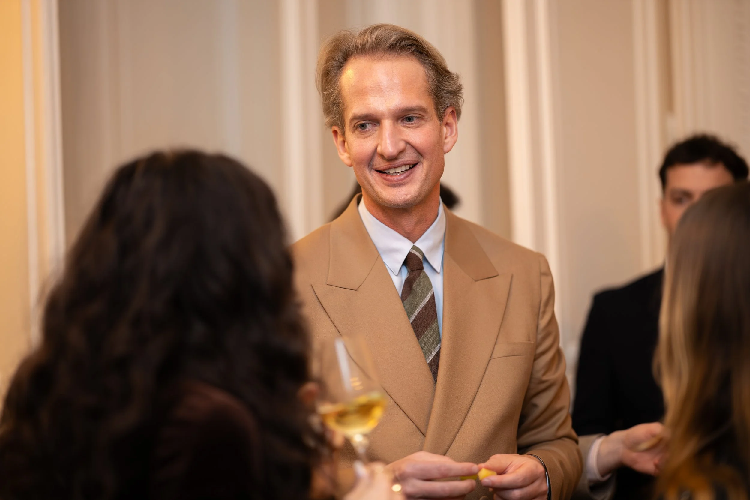 Un homme souriant en costume beige parlant à des personnes lors d'une réception ou d'une fête