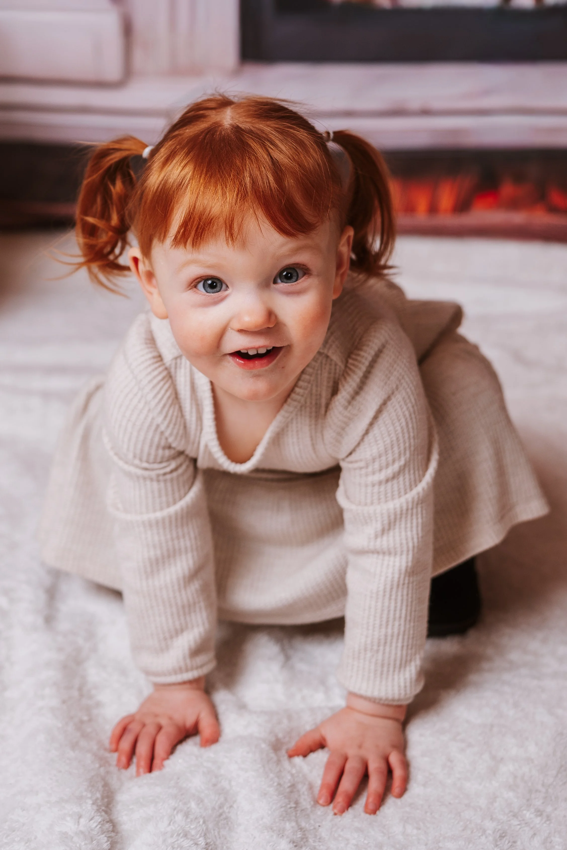 A young girl with red hair and blue eyes crawling on a white carpet, smiling and looking at the camera.