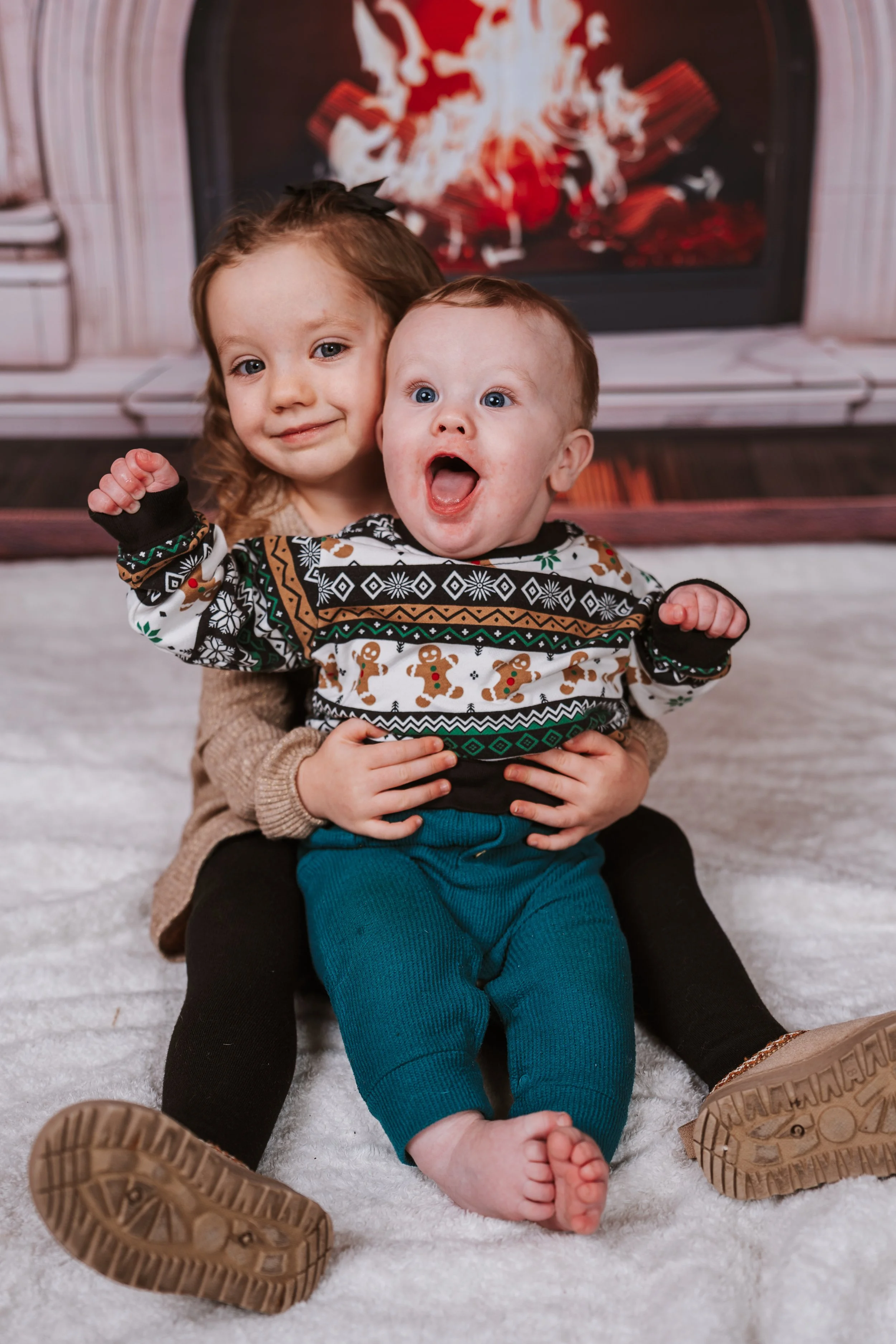 A young girl and a baby boy sitting in front of a fireplace with a roaring fire. The girl is hugging the boy, who has an excited expression with his mouth open. The girl has a small black bow in her hair and is smiling at the camera. The baby is wearing a festive holiday sweater with gingerbread men and Christmas patterns.