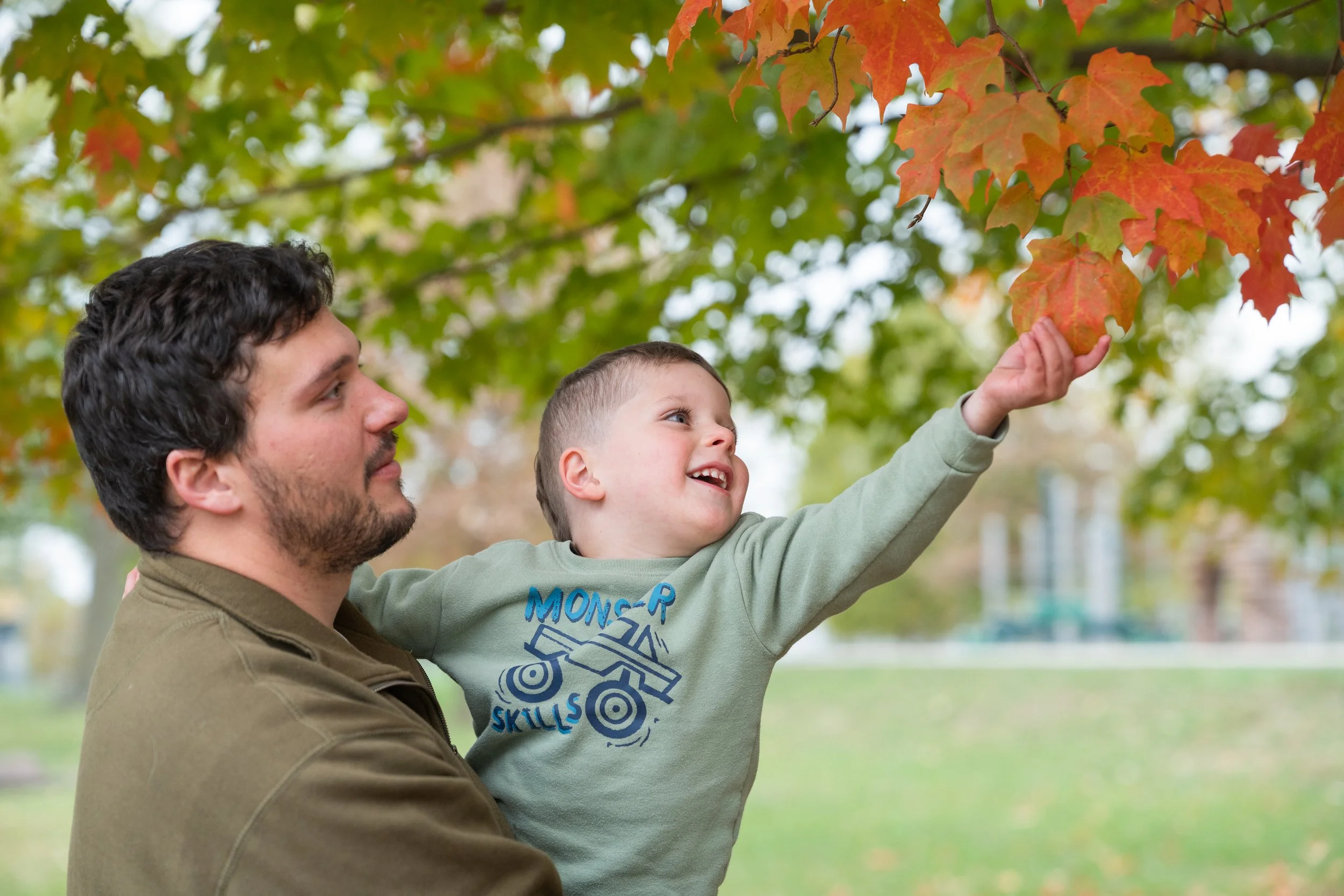 A man and young boy outdoors in a park during fall, with the boy reaching up to touch colorful orange and red autumn leaves on a tree.