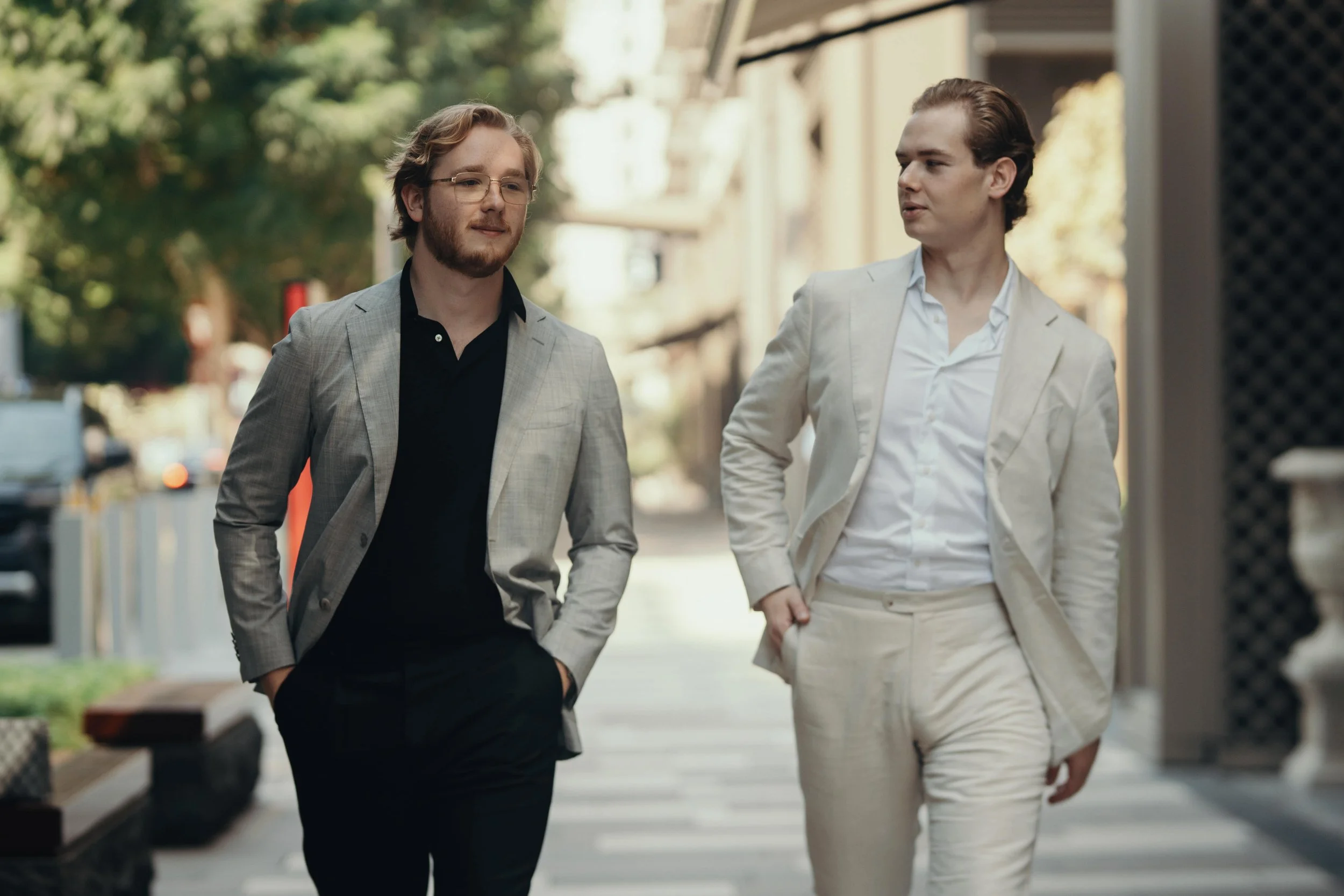 Two men in suits walking down a city street, one wearing a gray suit with a black shirt, the other in a light beige suit with a white shirt, trees in the background.