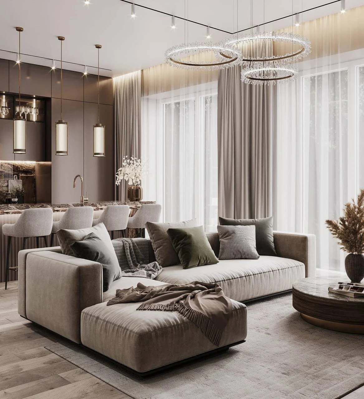 Modern living room with a beige sectional sofa, pillows, and a throw blanket. A round coffee table with decorative plants is in front. The dining area has bar stools and pendant lighting over a marble countertop. Large windows with sheer curtains illuminate the space.
