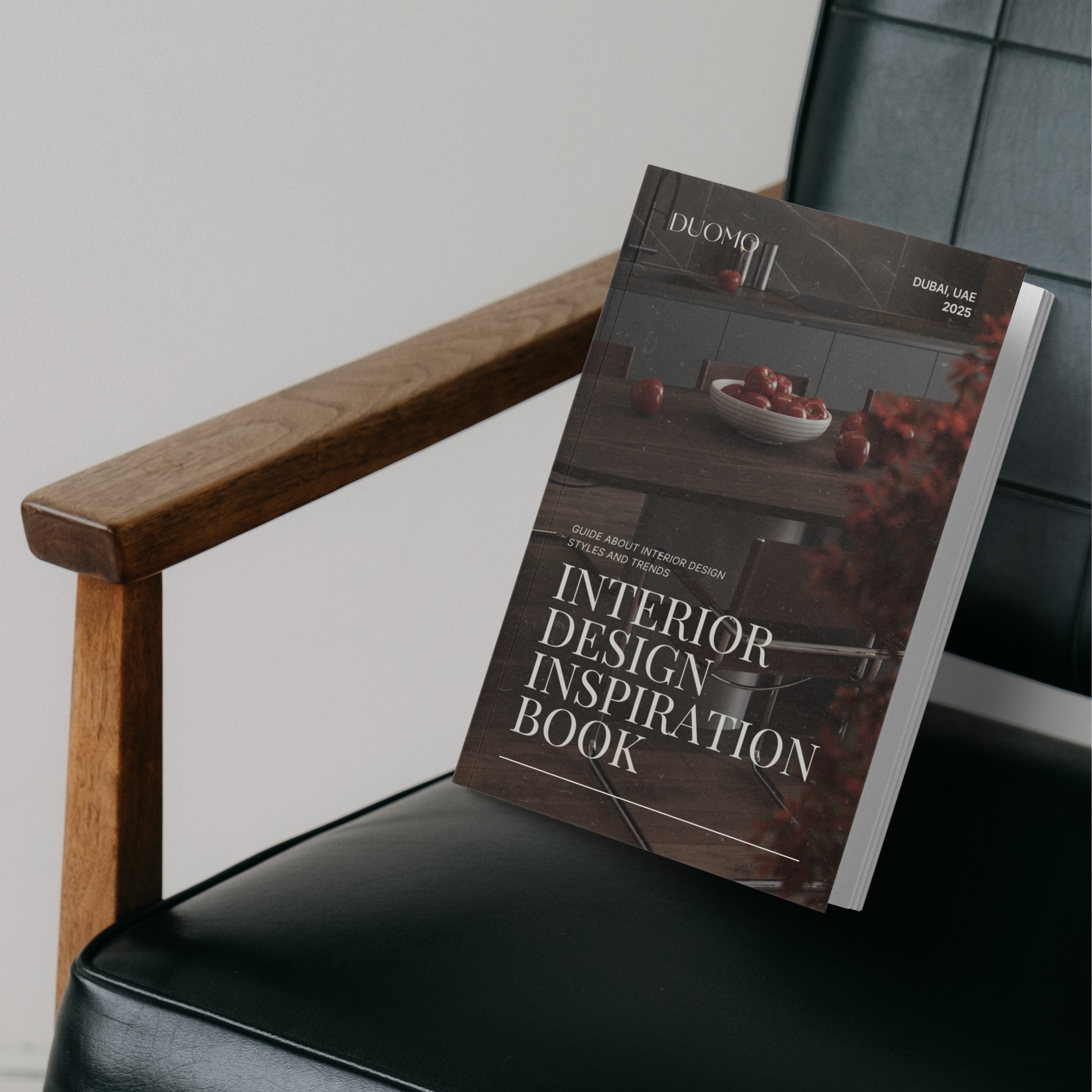 A book titled 'Interior Design Inspiration Book' resting on a black leather chair with a wooden armrest. The book cover features a kitchen countertop with red apples and kitchen decor. Published by DUOMO, dated 2025 in Dubai, UAE.