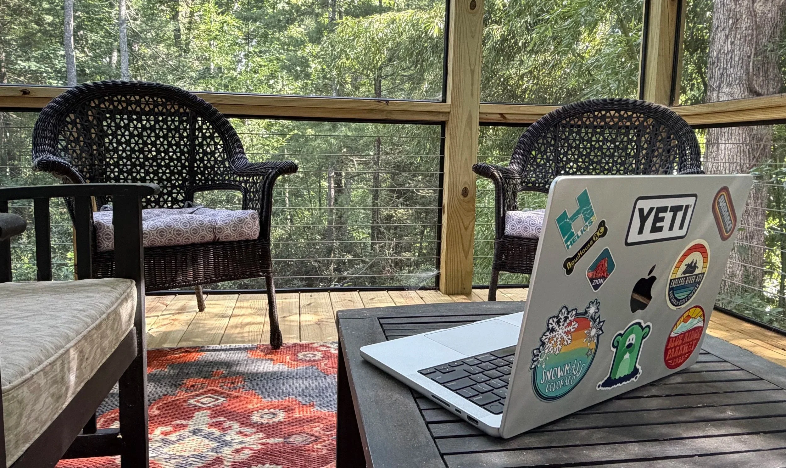 Laptop covered in outdoor brand stickers on a table on a screened porch overlooking the woods — David Kreeger's writing spot for GoPedal's Musings posts.