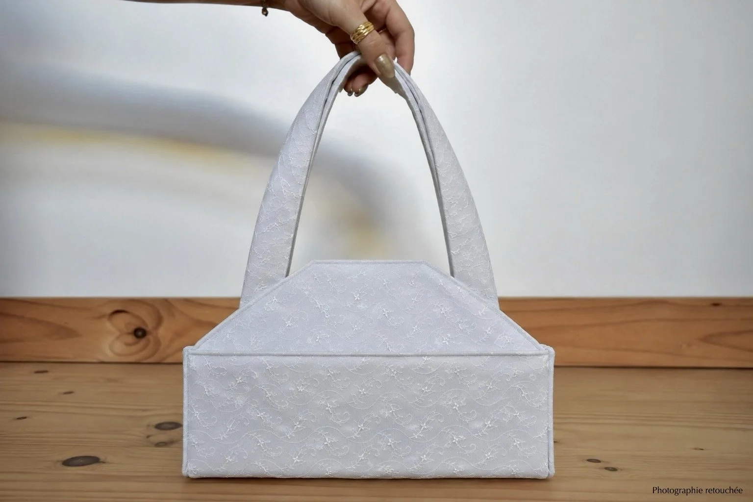 Une main tenant un sac à main de couleur blanc avec des poignées, posé sur une surface en bois.