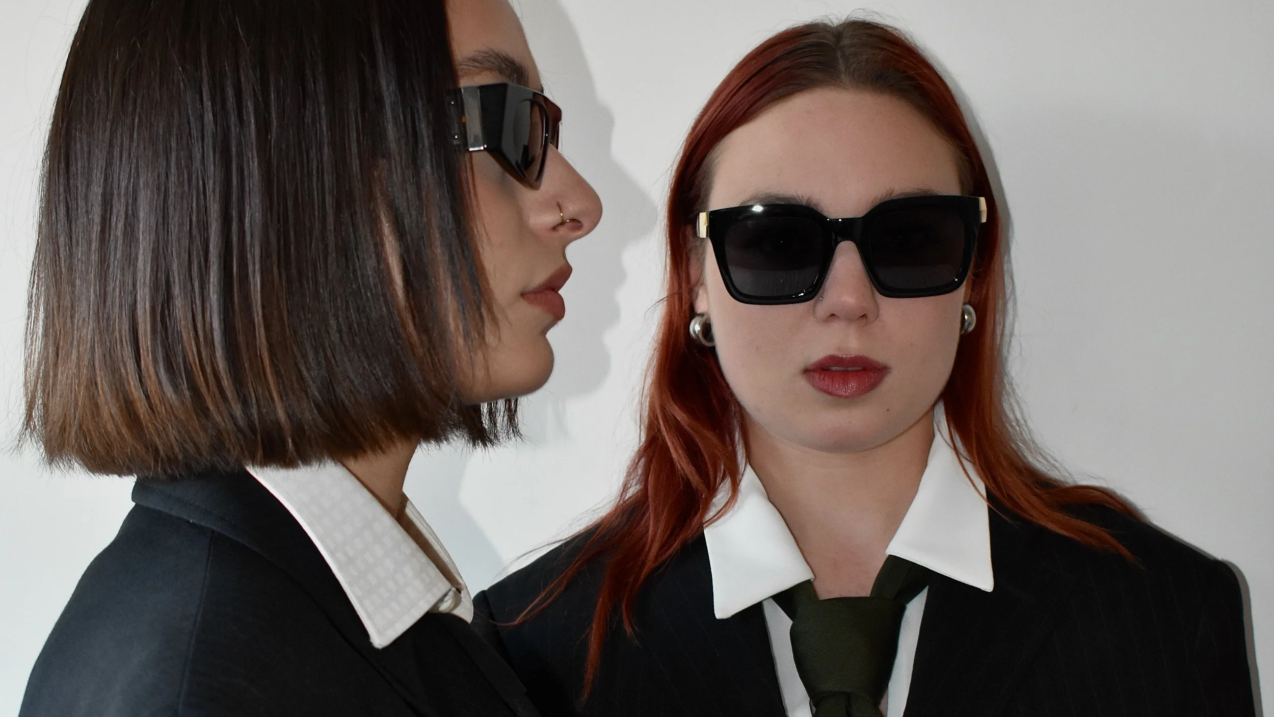 Deux femmes portant des costumes noirs et des lunettes de soleil, l'une de face et l'autre de profil, se trouvant contre un fond blanc.