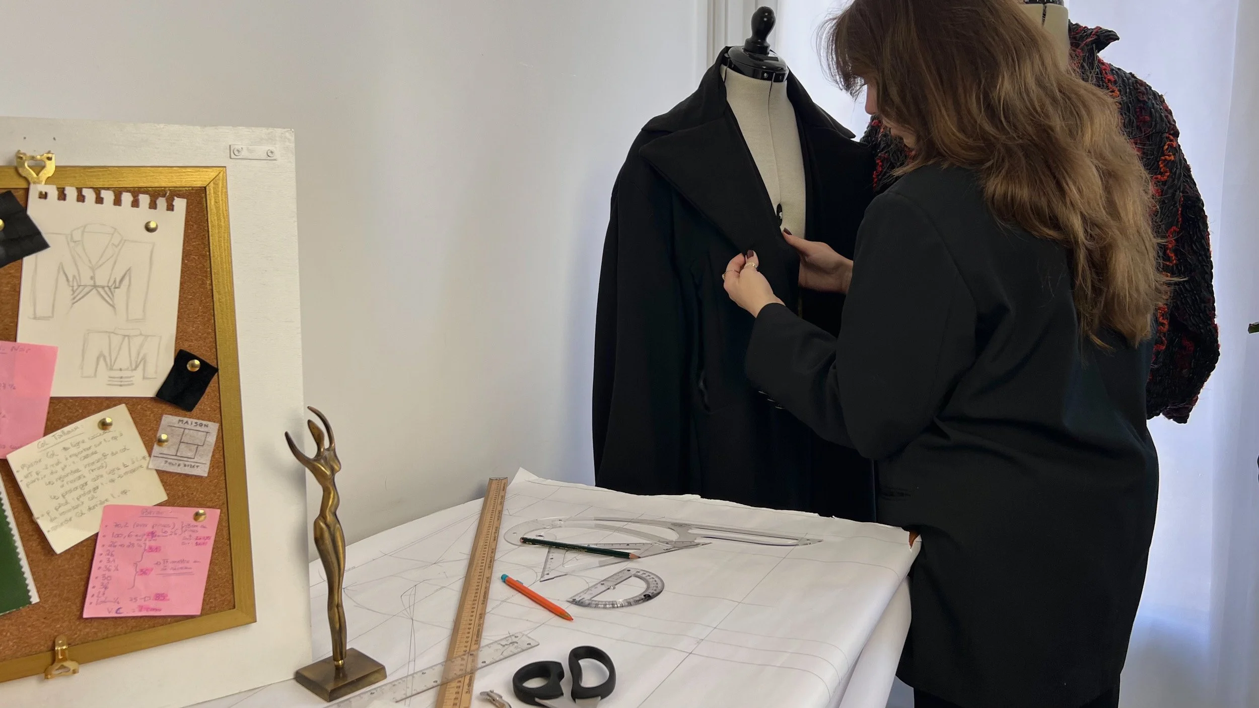 Une femme travaille sur un mannequin en examinant un vêtement noir. La scène se passe dans un espace de travail dédié à la couture ou au design de mode, avec des outils, des plans et un tableau d'idées ou de croquis à proximité.