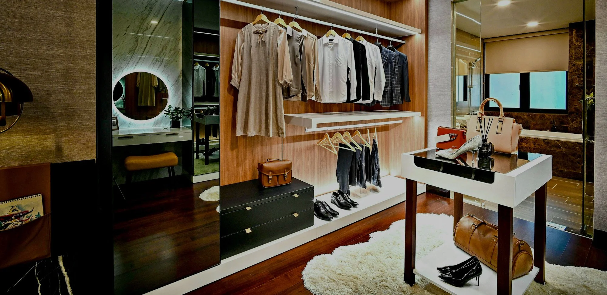 Une étagère de vêtements dans un dressing, avec des chemises, des pantalons et des accessoires comme des sacs, une lampe, un miroir, et un fauteuil visible en arrière-plan.