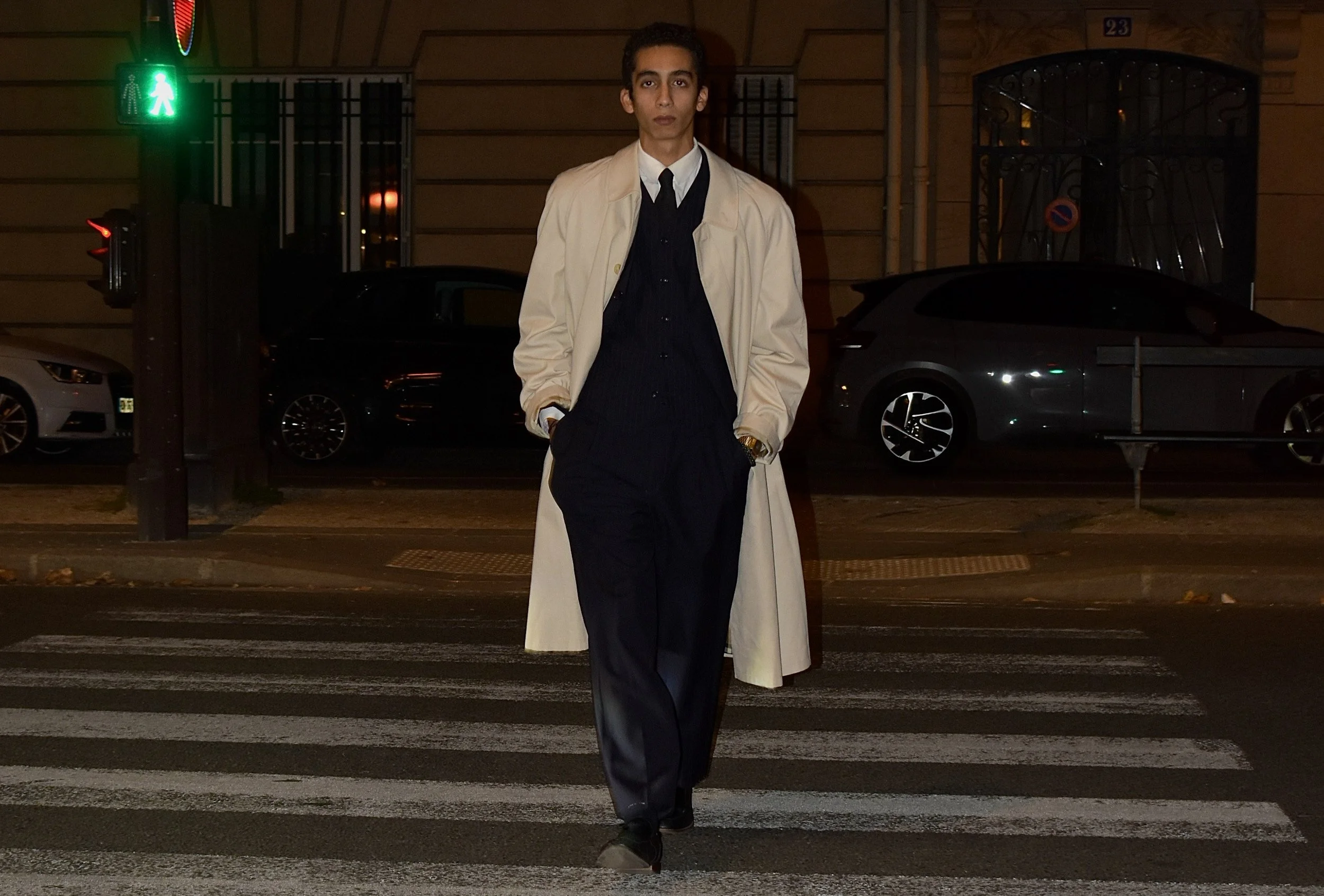 Homme debout sur un passage pour piétons, portant un trench beige, une chemise blanche, un costume bleu foncé, la nuit, avec des voitures en arrière-plan.