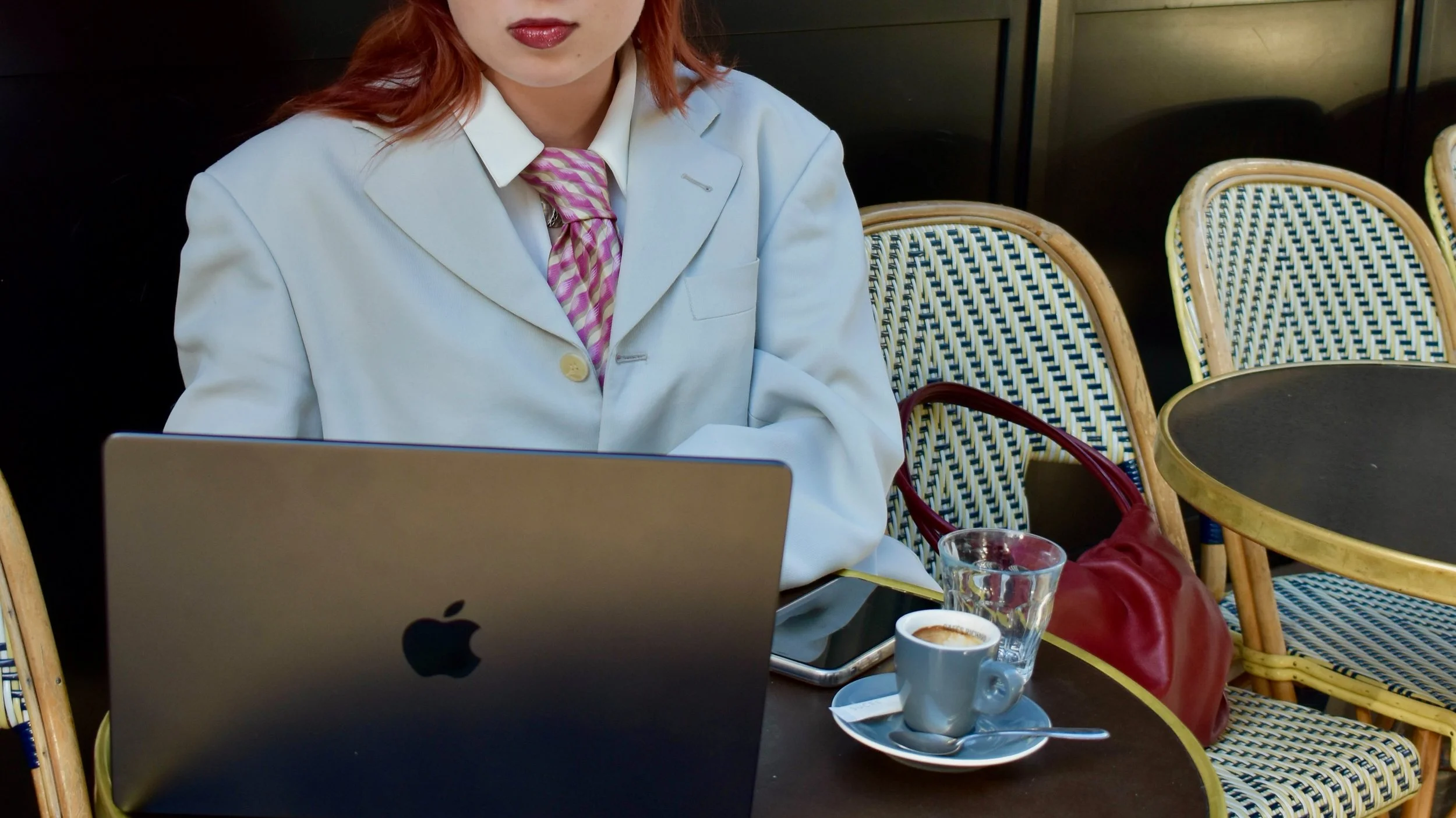 Une femme portant une veste grise, assise à une table avec un ordinateur portable Apple, un verre d'eau, une tasse de café, et un sac rouge, en arrière-plan des chaises en rotin et un mur sombre.