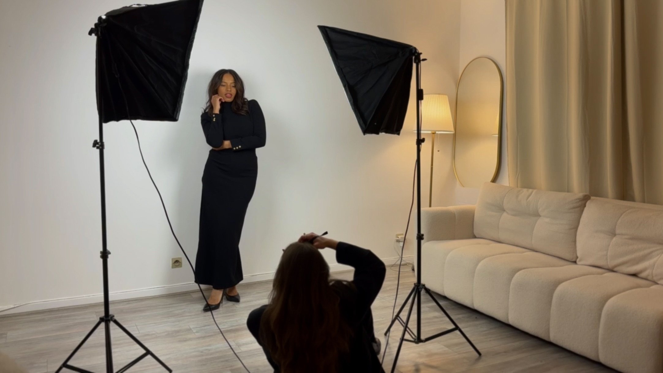 Une femme habillée en noir pose lors d'une séance photo à l'intérieur, avec deux softboxes qui l'éclairent, un miroir accroché au mur, un sofa beige et une lampe dans une pièce chaleureuse.
