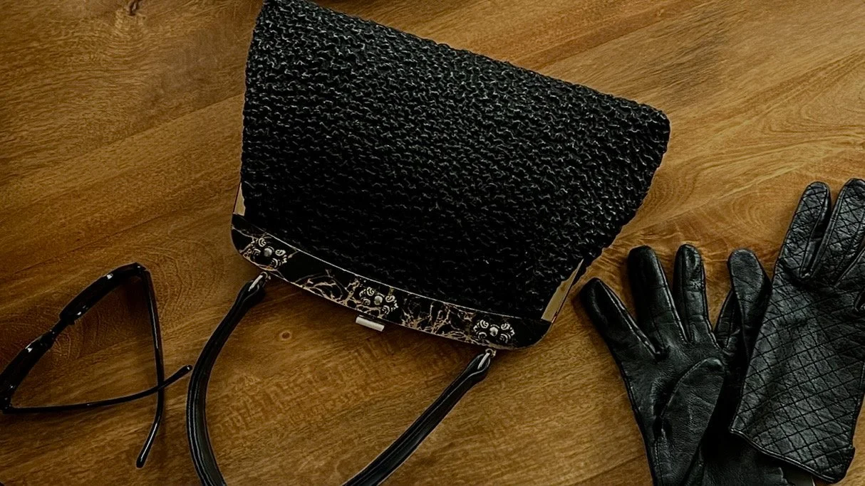 Une paire de lunettes, un sac à main noir en simili cuir avec une texture bouclée, et une paire de gants en cuir noir avec un motif en croix, posés sur une table en bois.