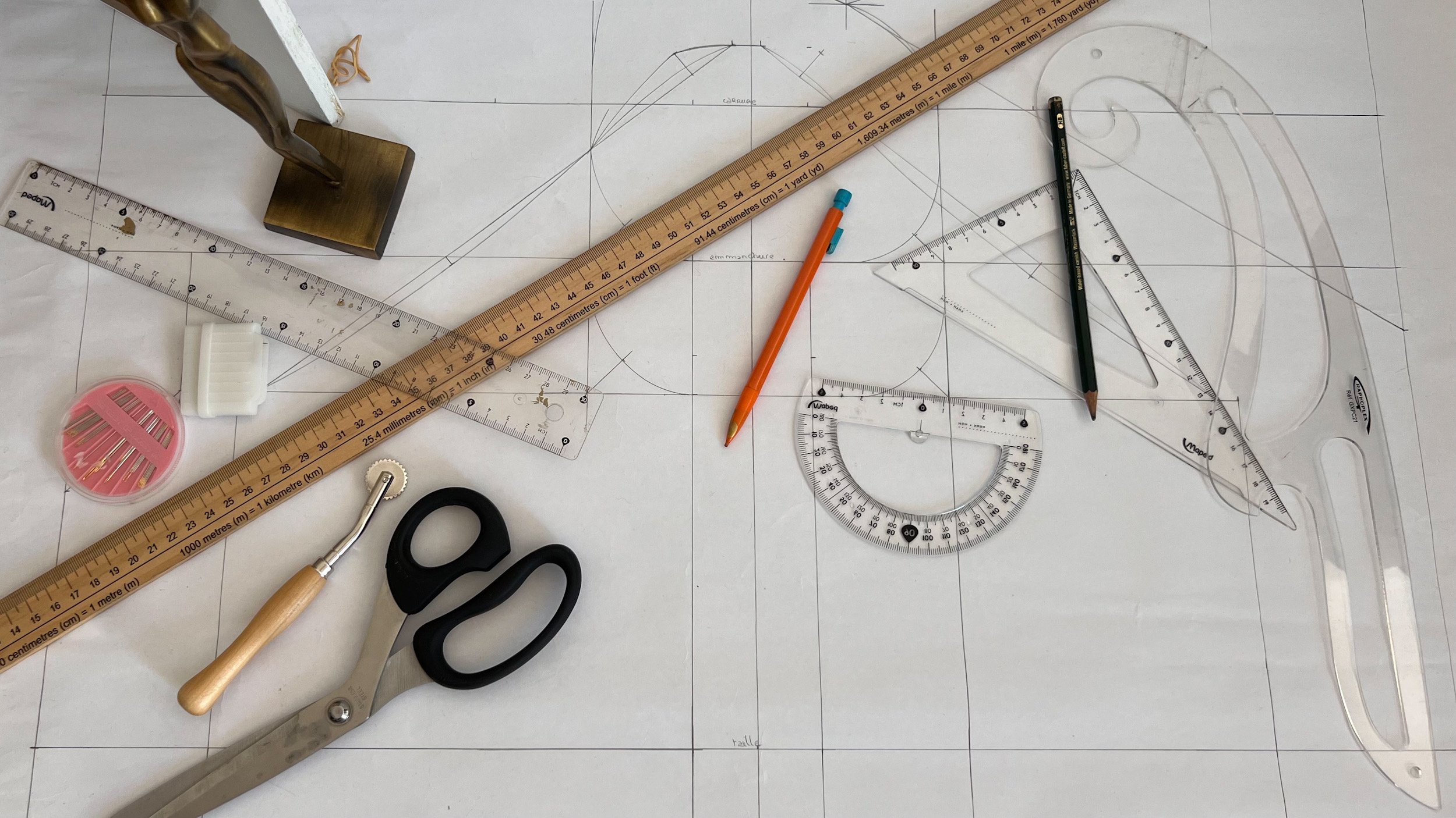 Plan de dessin technique avec outils de dessin comme règle, compas, équerre, stylo mécanique, ciseaux, encre, et matériel de couture sur une surface blanche.