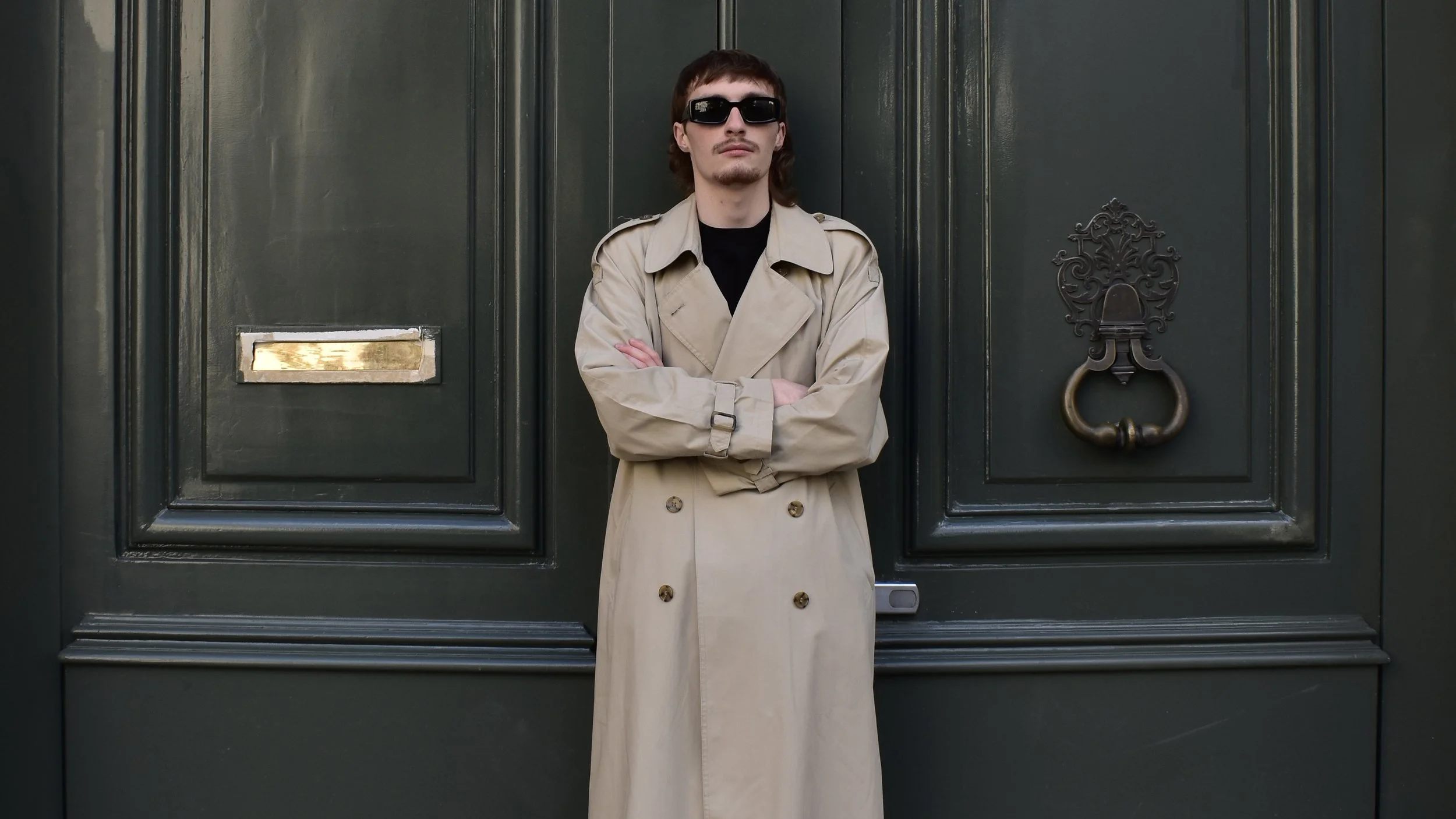 Un jeune homme portant des lunettes de soleil, une trench beige, se tient croisé devant une grande porte en bois vert foncé.