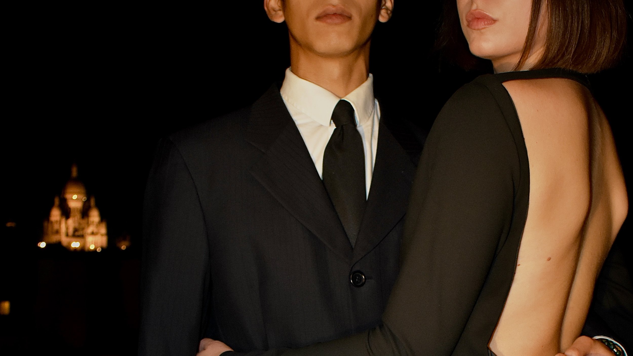 Deux personnes habillées élégamment lors d'une soirée nocturne avec la ville en arrière-plan. La personne à gauche porte un costume noir avec une chemise blanche et une cravate noire, et la personne à droite porte une robe noire avec un dos nu.