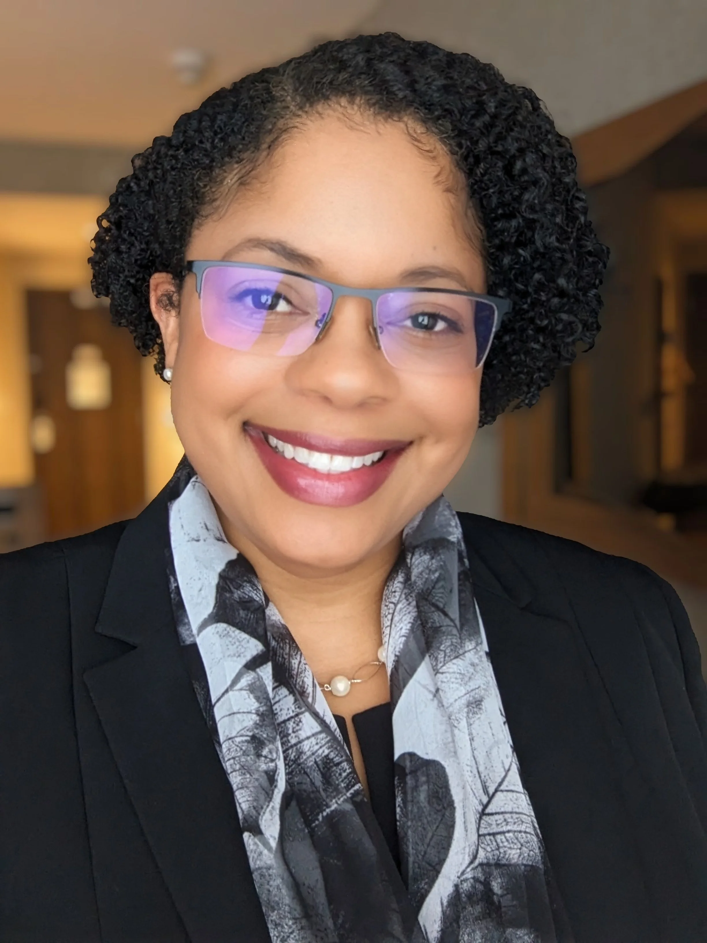 A smiling woman with short, curly black hair wearing glasses, a black blazer, a patterned blouse, and a pearl necklace inside a warmly lit room.