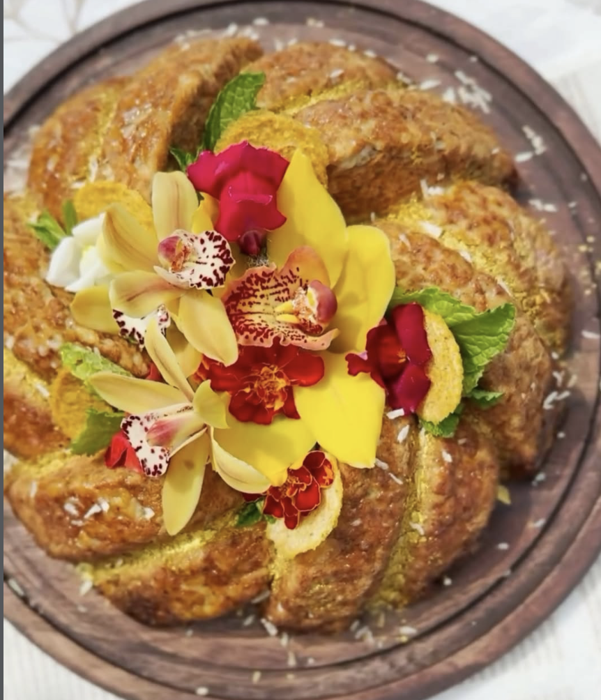 A round, golden brown bread pudding garnished with edible flowers and fresh mint leaves on a wooden plate.