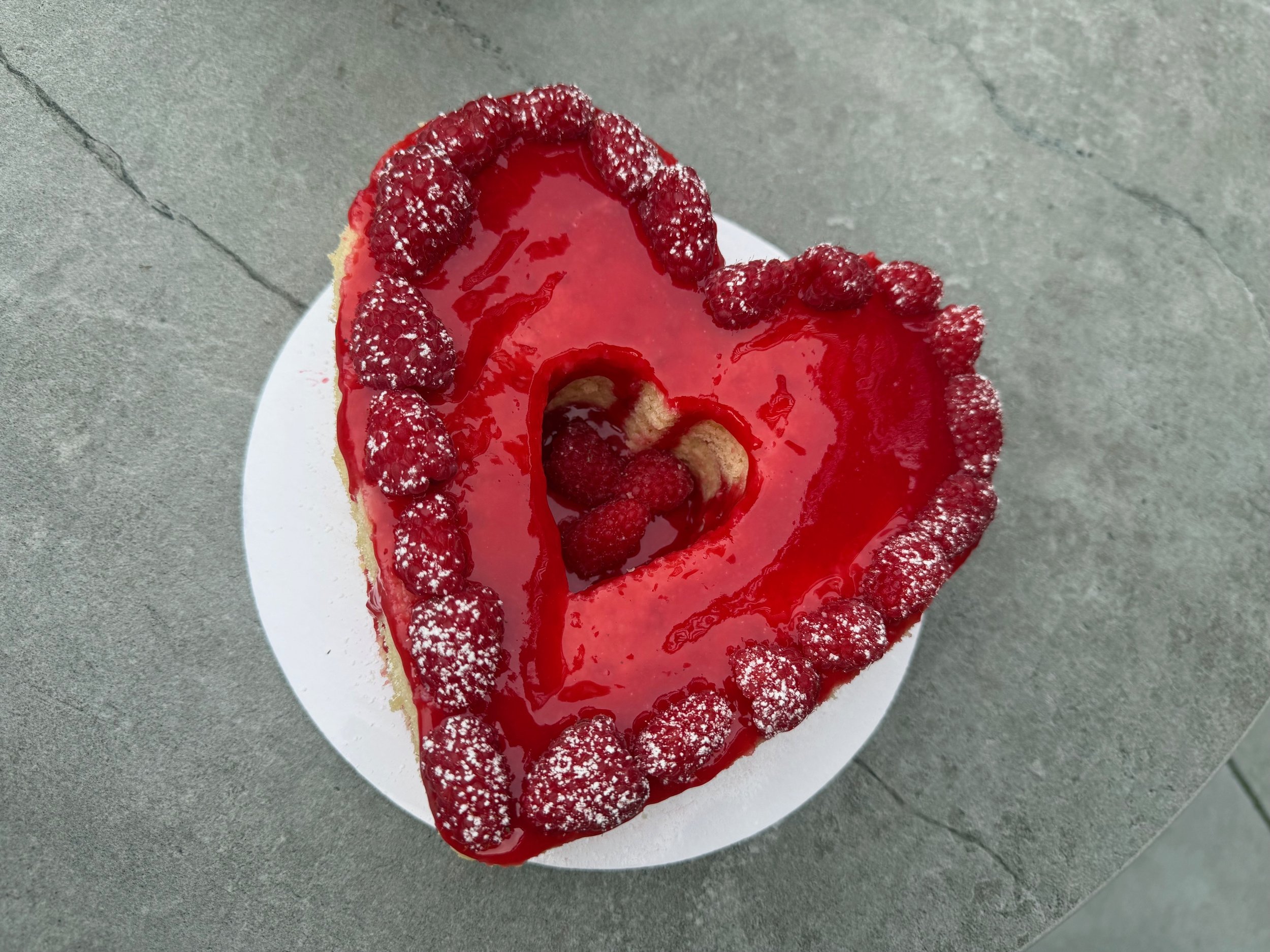 A slice of cake topped with red fruit glaze and blackberries on the border, served on a white plate on a gray surface.